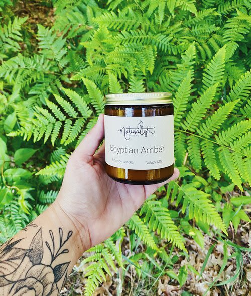 Hand holding an amber jar candle labeled Egyptian Amber in front of bright green ferns.