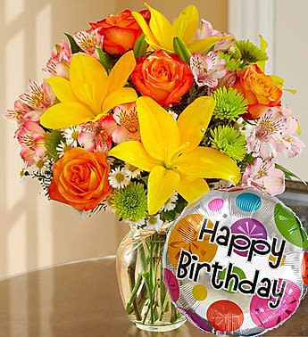 Bouquet of yellow lilies, orange roses, and pink alstroemeria in a glass vase with a birthday balloon.