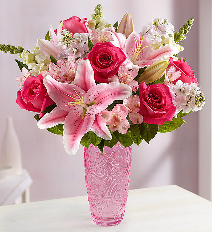 Arrangement of pink lilies and roses with white accent flowers in a tall pink glass vase