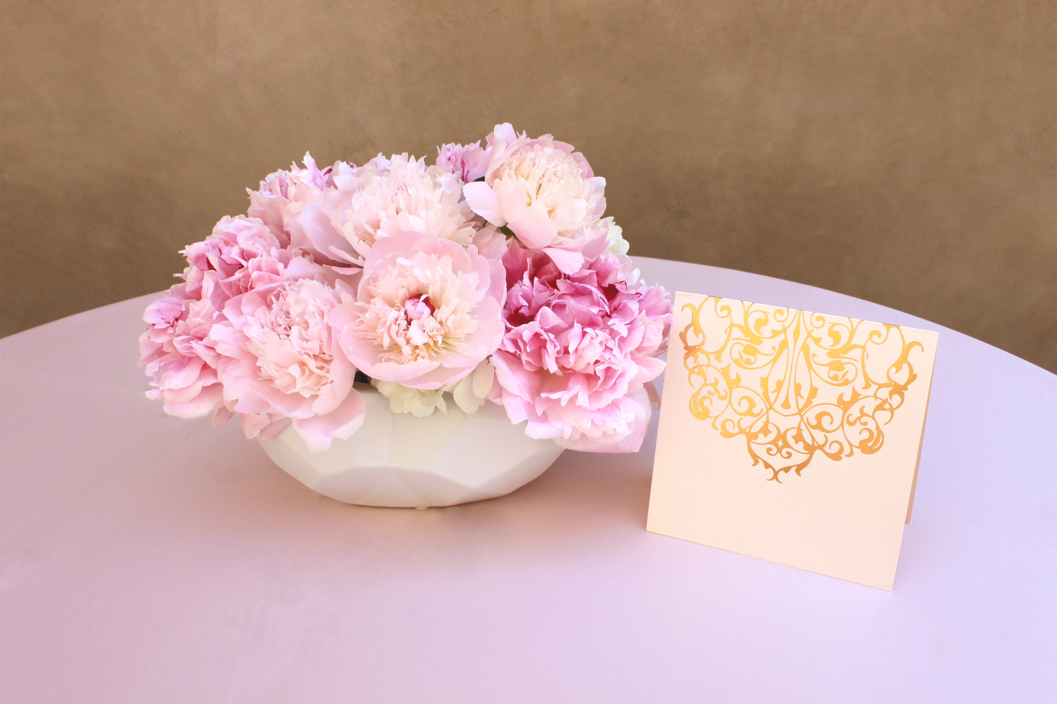 Pink peonies in a white bowl with a decorative card beside them
