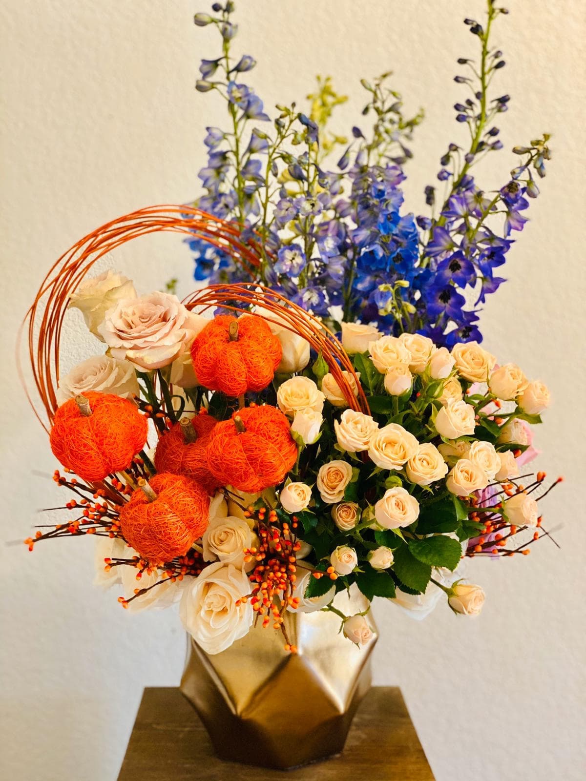 Bouquet of cream roses and blue delphinium with orange pumpkin accents in a gold vase