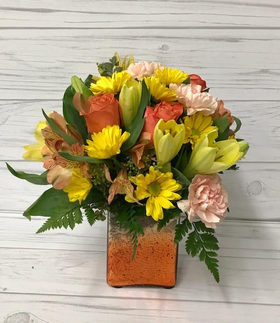 Mixed bouquet of yellow, peach, and coral flowers in a square glass vase