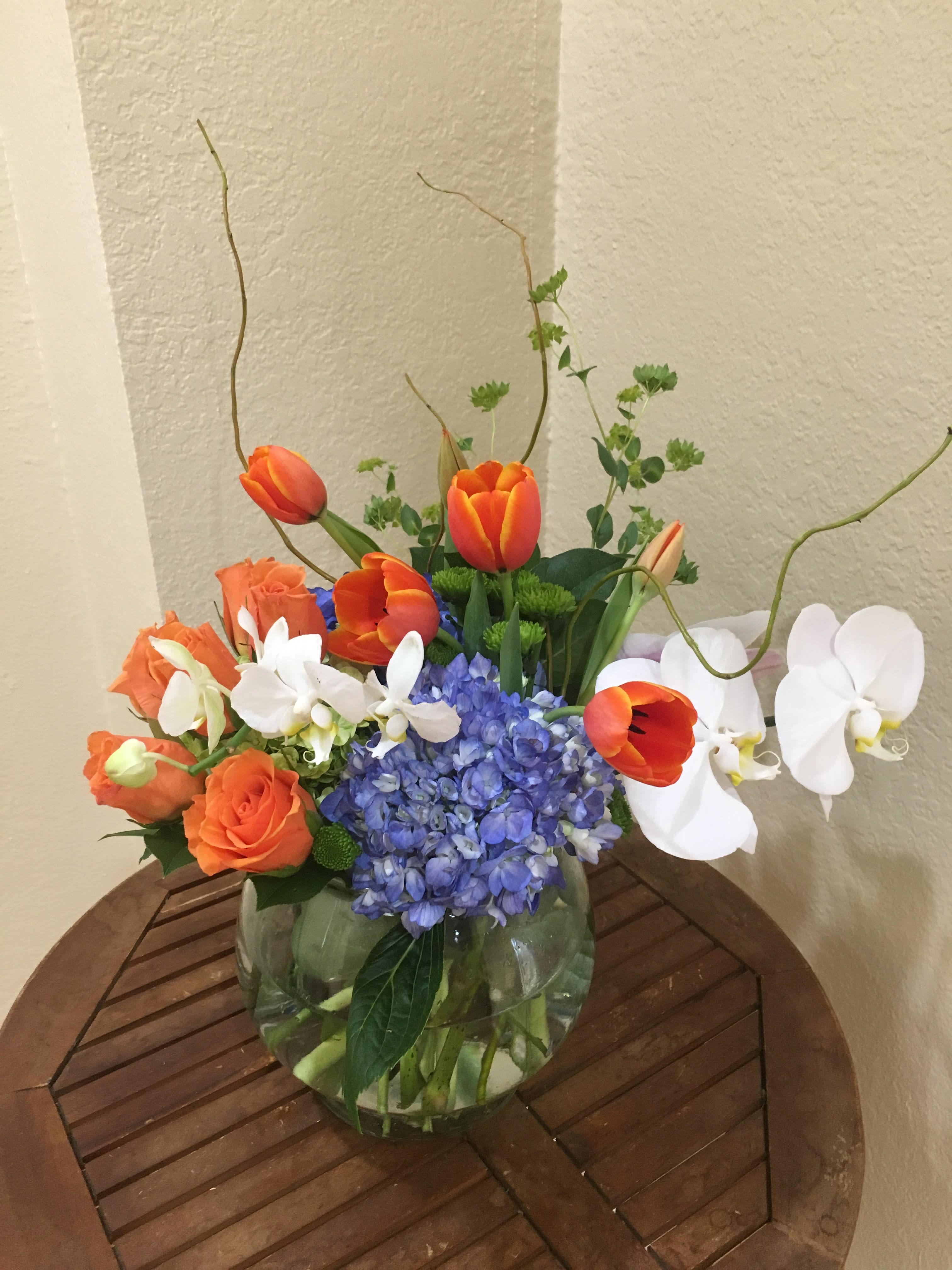 Orange tulips, roses, white orchids, and blue hydrangea in a glass vase