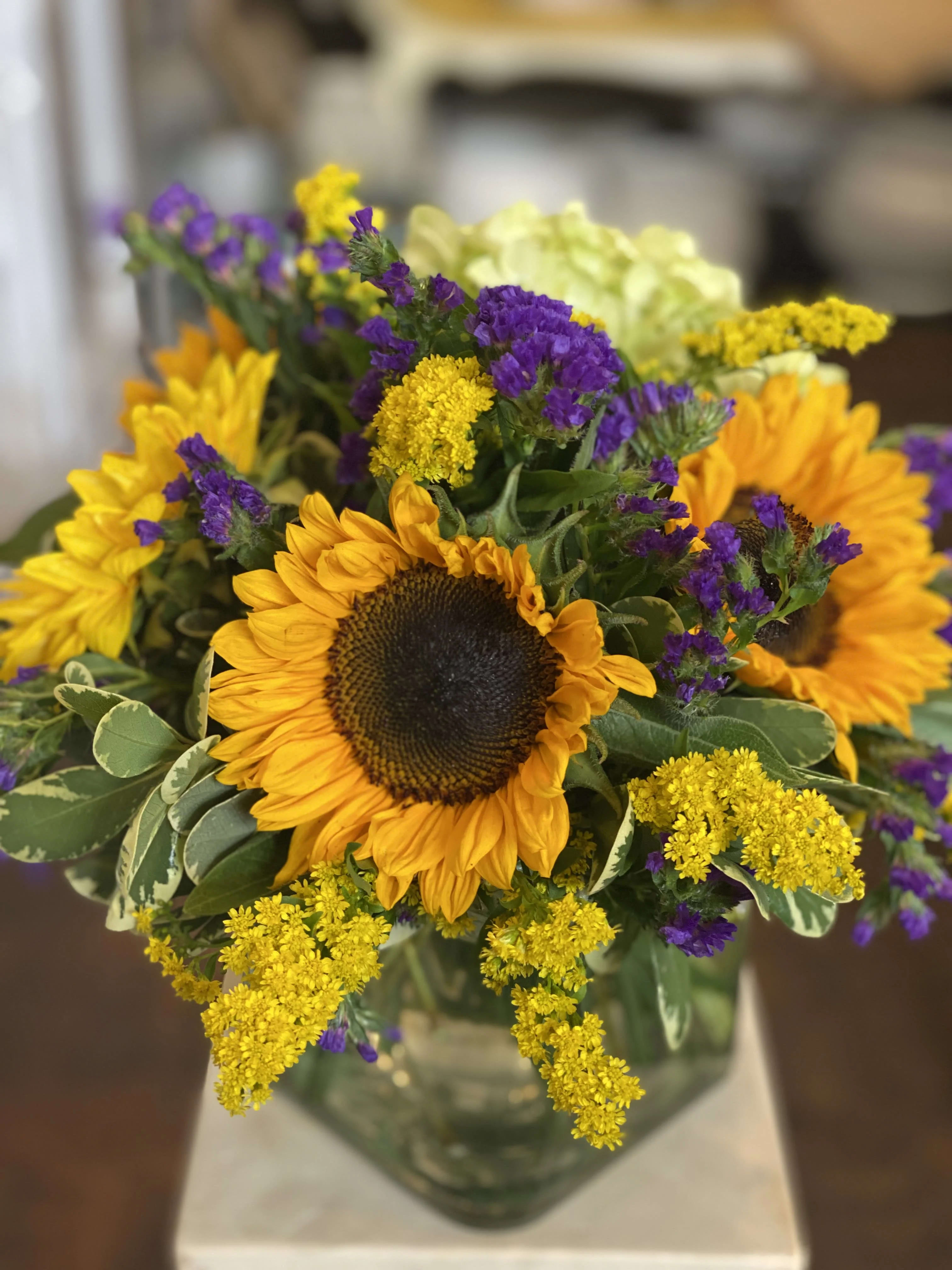 Golden Sunflower  - These beautiful sunflowers are prominently displayed among an arrangement of luscious greens, shining bright and looking fresh. Green hydrangeas, purple statice, surround the sunflowers, all displayed in a clear 4"x4" cube vase.