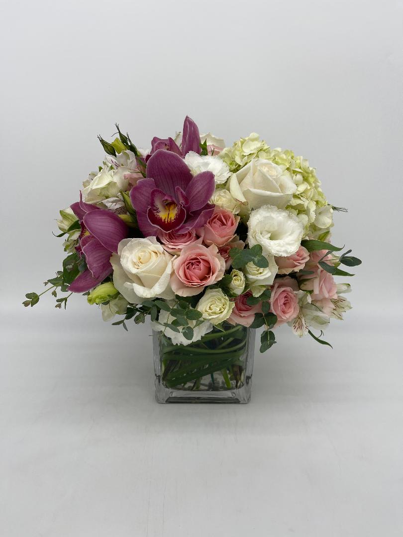 Compact arrangement of pink and white roses with purple orchids in a clear glass cube vase