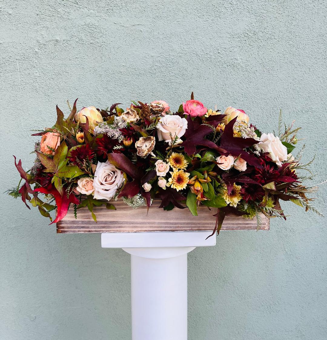 Low rectangular arrangement of blush roses and autumn-toned flowers in a wooden box on a white pedestal.