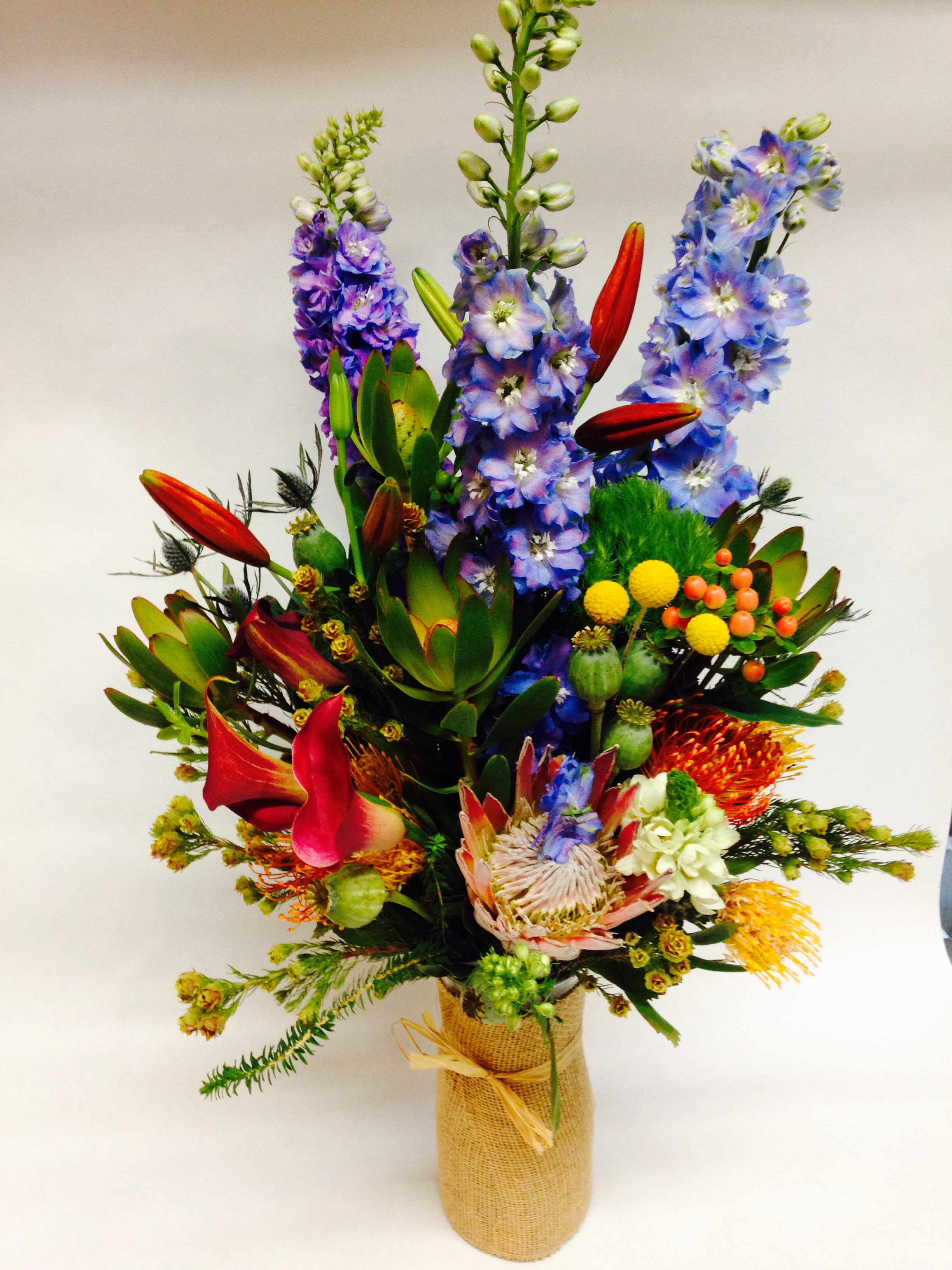 Tall mixed bouquet with blue-purple flowers and red lilies in a burlap-wrapped vase