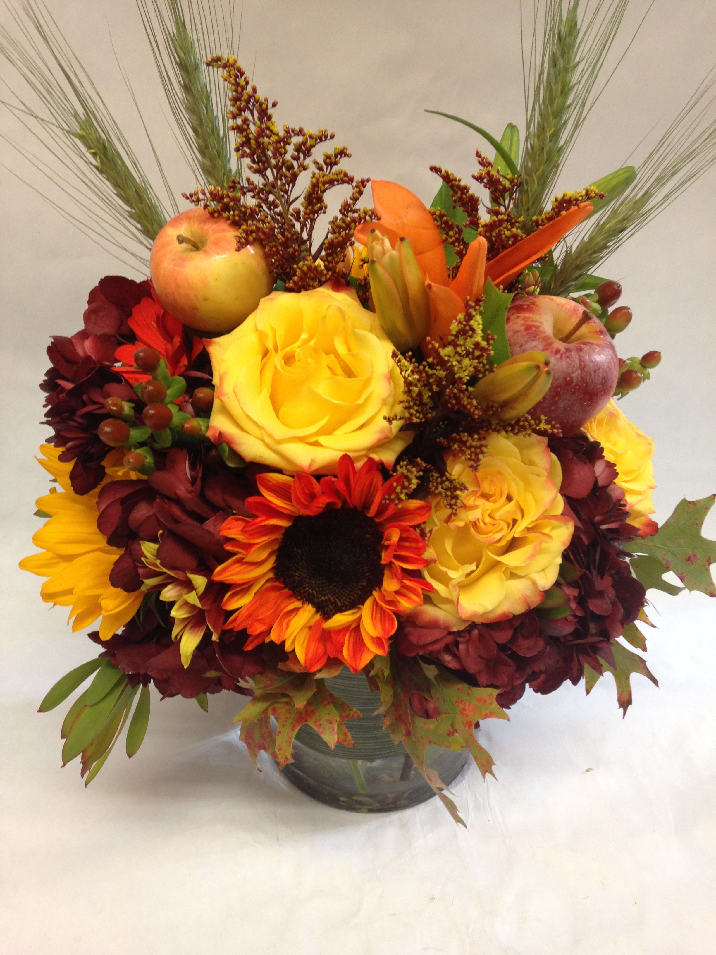 Mixed autumn bouquet with roses, sunflowers, apples, and lilies in a glass vase