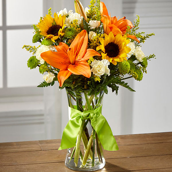 Bouquet of orange lilies, sunflowers, and white carnations in a glass vase with a green ribbon