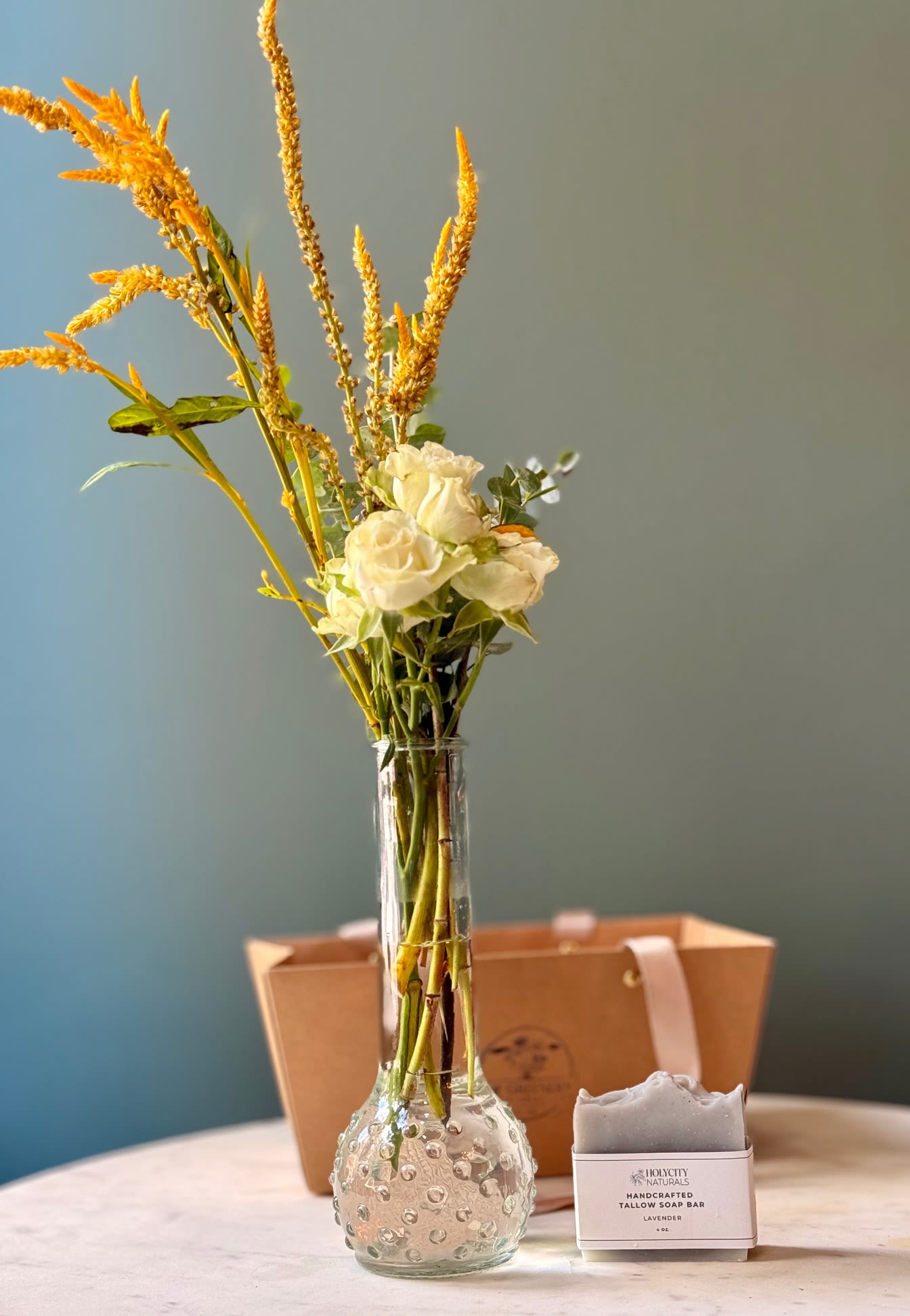 White roses in a clear glass vase with a soap bar gift box beside it.