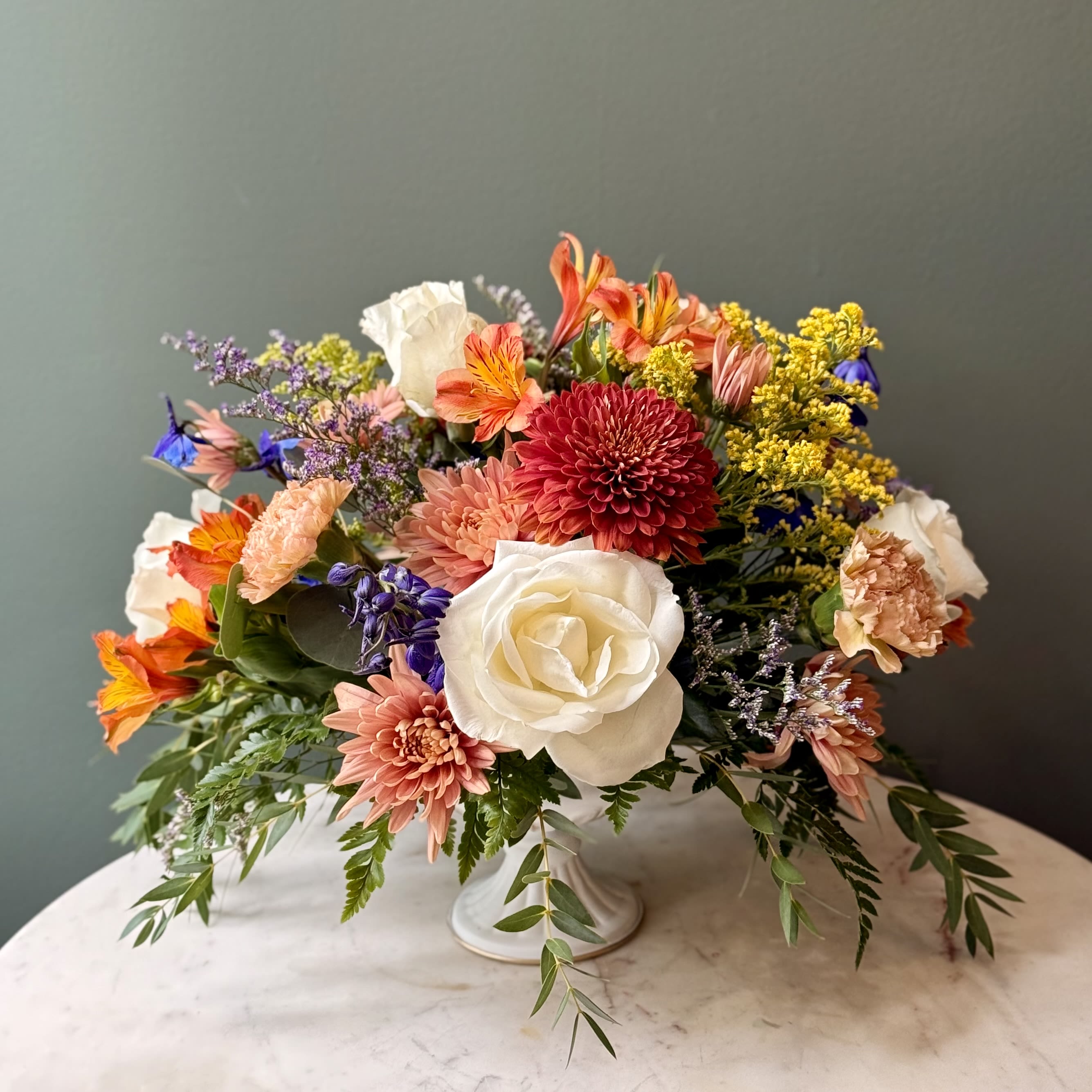 Autumn Blooms - A soft, sophisticated fall floral design for moments that deserve a touch of elegance. Featuring: - Ecuadorian Roses - Alstroemeria - Delphinium - Solidago - Cremone - Cushion Mums - Styled with premium seasonal foliage Presentation Options: White urn Every order includes: A custom message card - simply enter your message at checkout and we'll take care of the rest. Please note: Flowers are subject to seasonal availability. If a listed flower is unavailable, our designers will substitute it with flowers of a similar shape, color, and style to maintain the overall aesthetic.