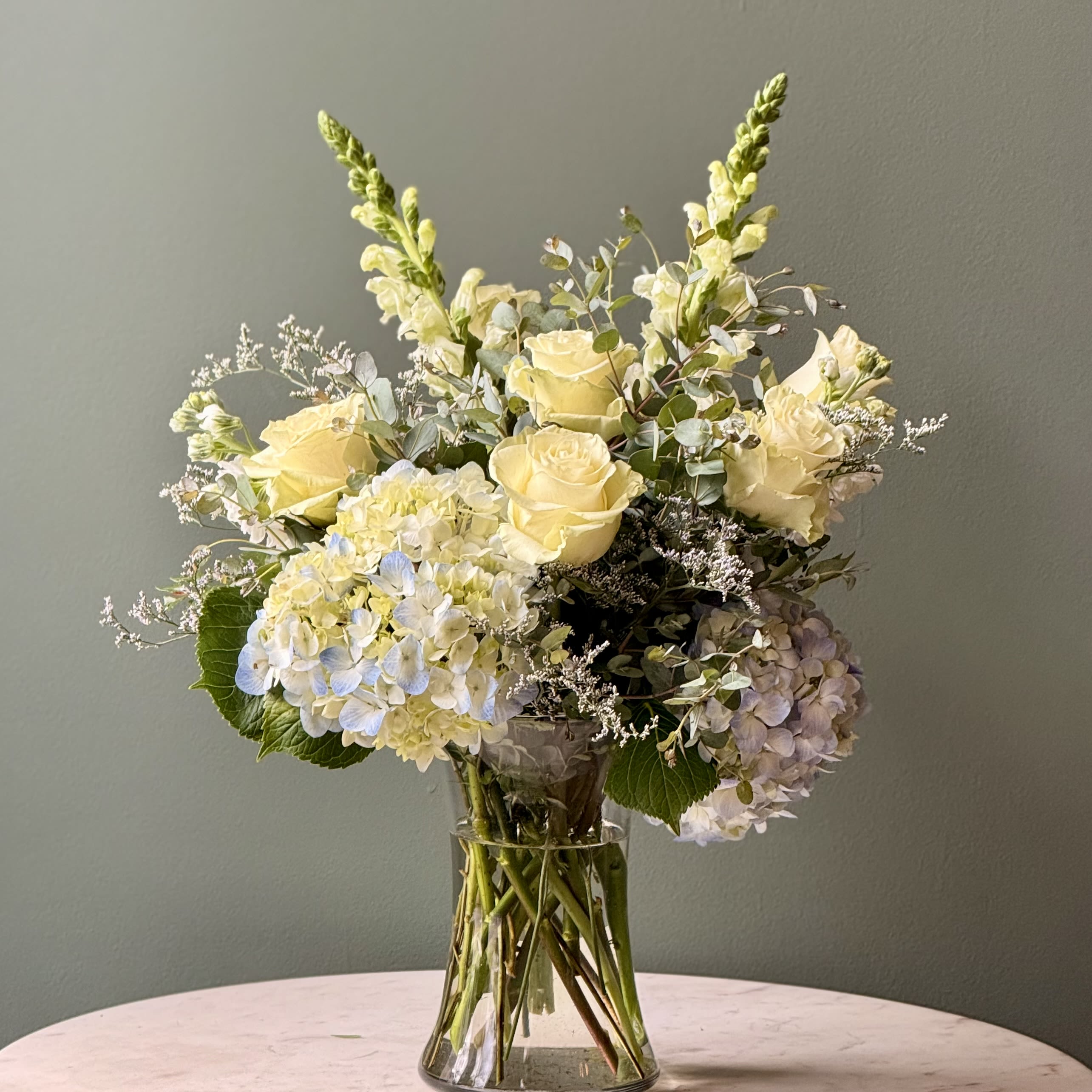Arrangement of pale yellow roses, white hydrangeas, and snapdragons in a clear glass vase.