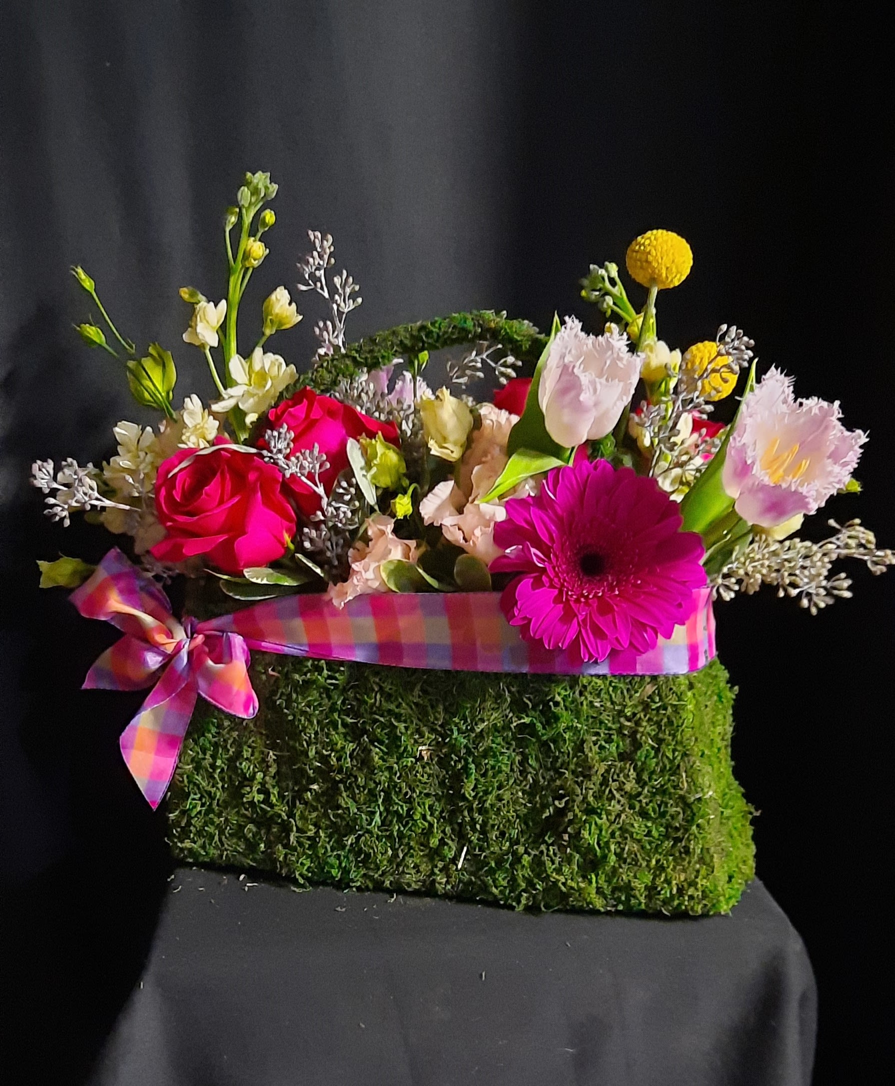 Bright mixed flowers in a moss-covered handbag container with a plaid ribbon bow