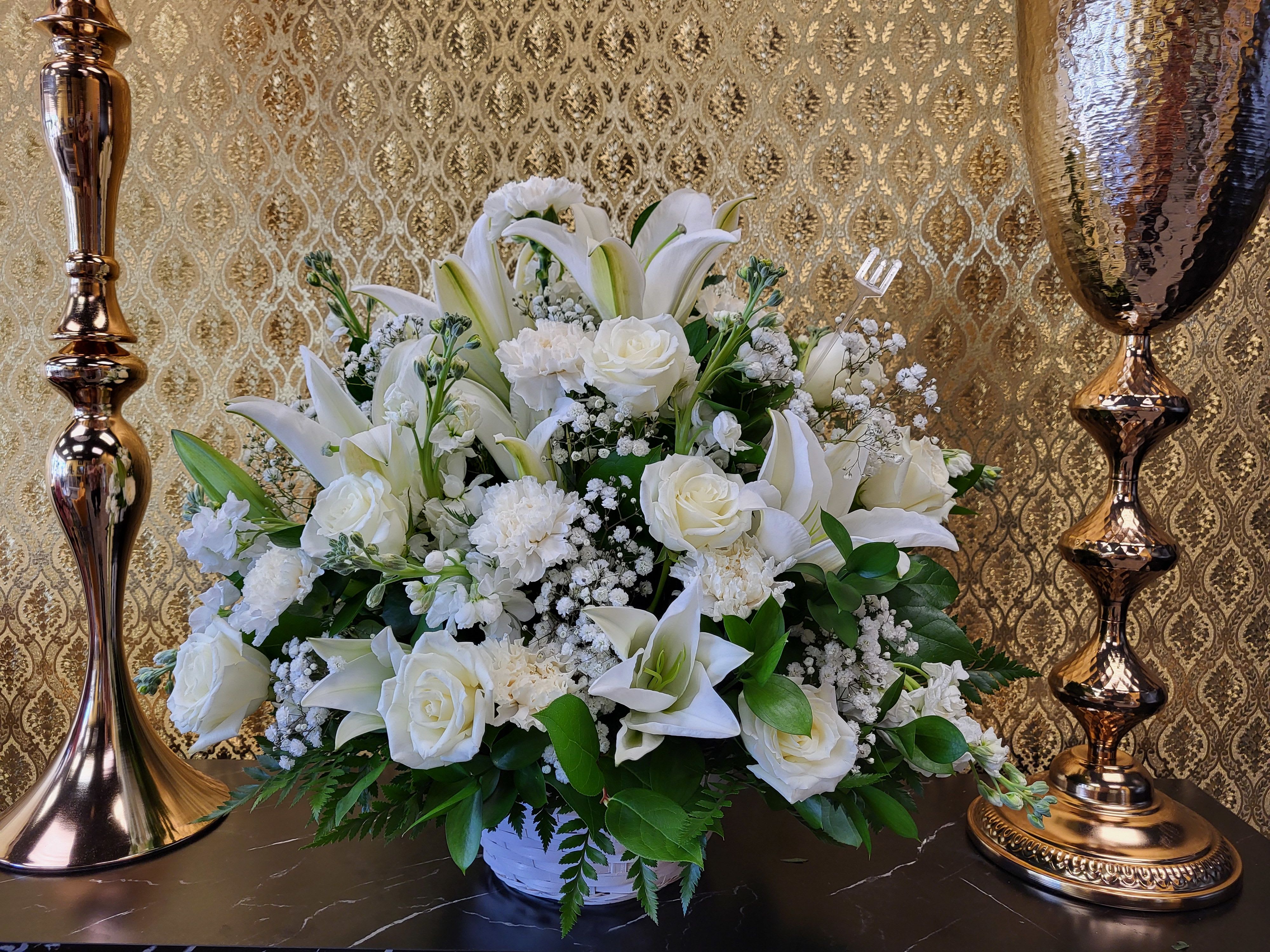 White lilies, roses, and carnations arranged in a white container with gold decor in the background.