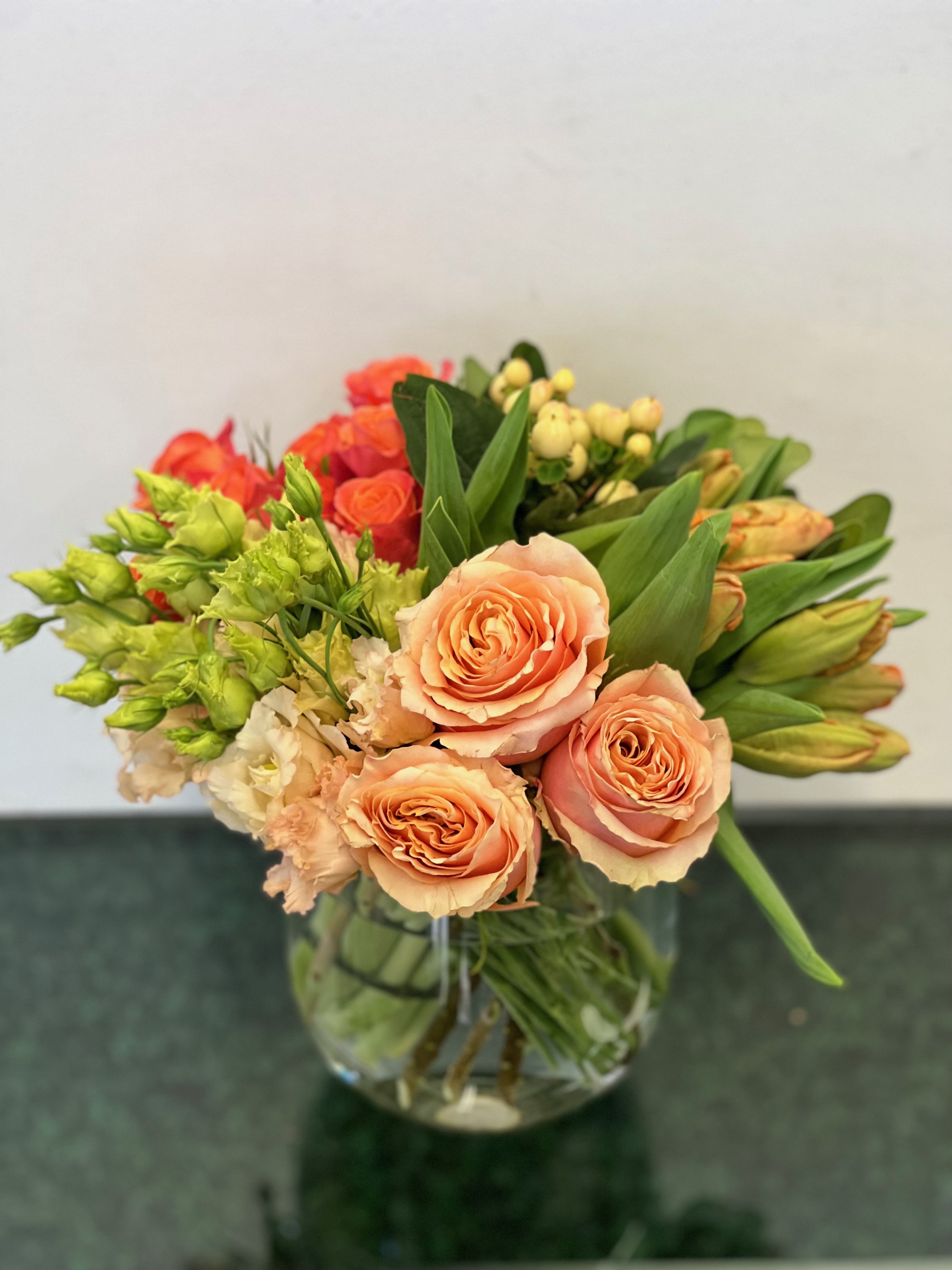 Tart and Juicy Apricots and Oranges - A tart arrangement of roses, tulips, spray roses lisianthks and hypericum in a simple glass vase.  *Please note that we are doing our best to replicate our designs exactly as shown, but with the  supply chain issues these days we will design as closely as possible.  If something is not available that will change the overall look of the arrangement we will reach out to let you know.