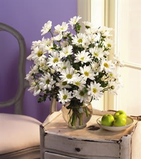 White daisy bouquet in a glass vase beside a bowl of green apples