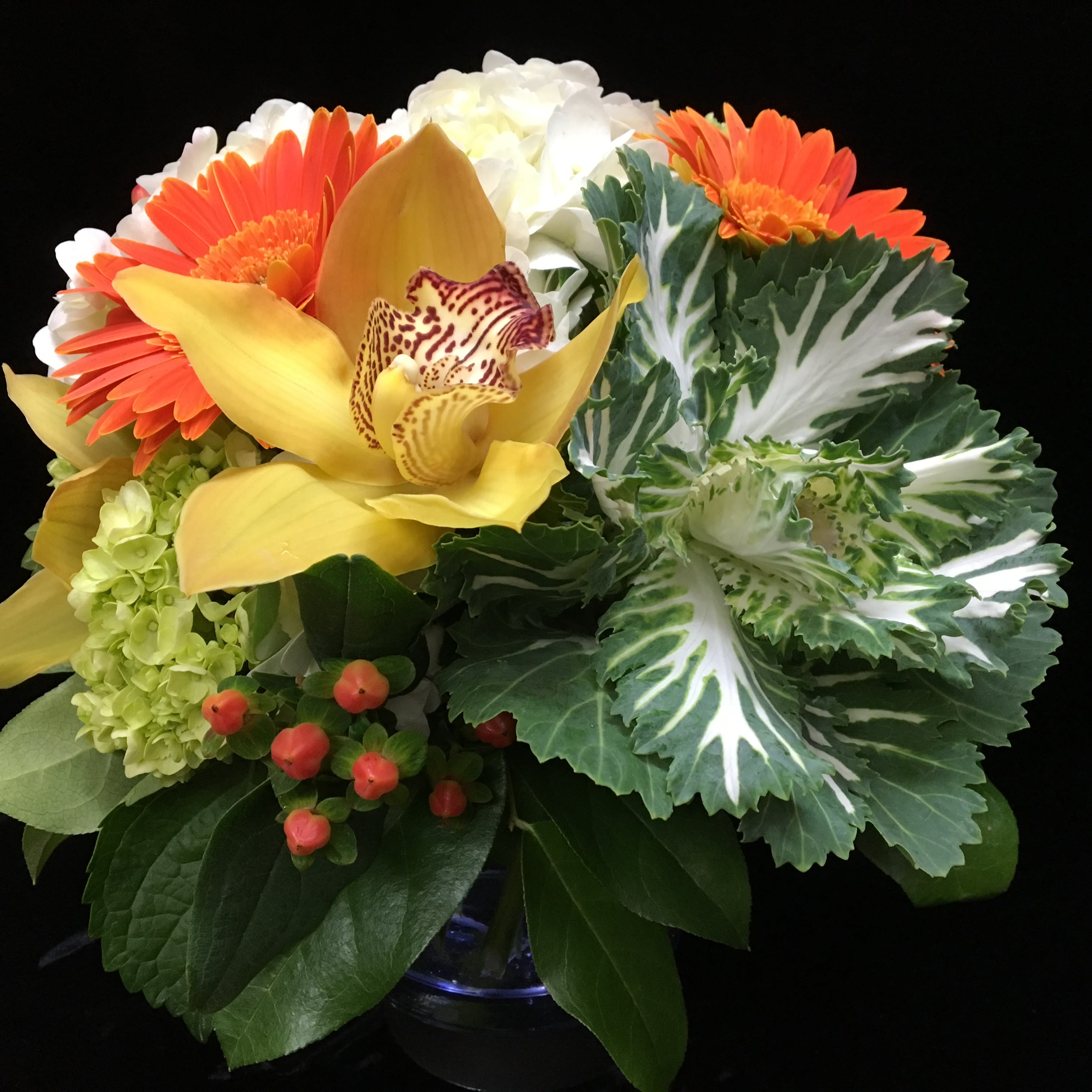 Compact arrangement of orange gerberas, yellow orchid, hydrangeas, and ornamental cabbage in a glass vase.
