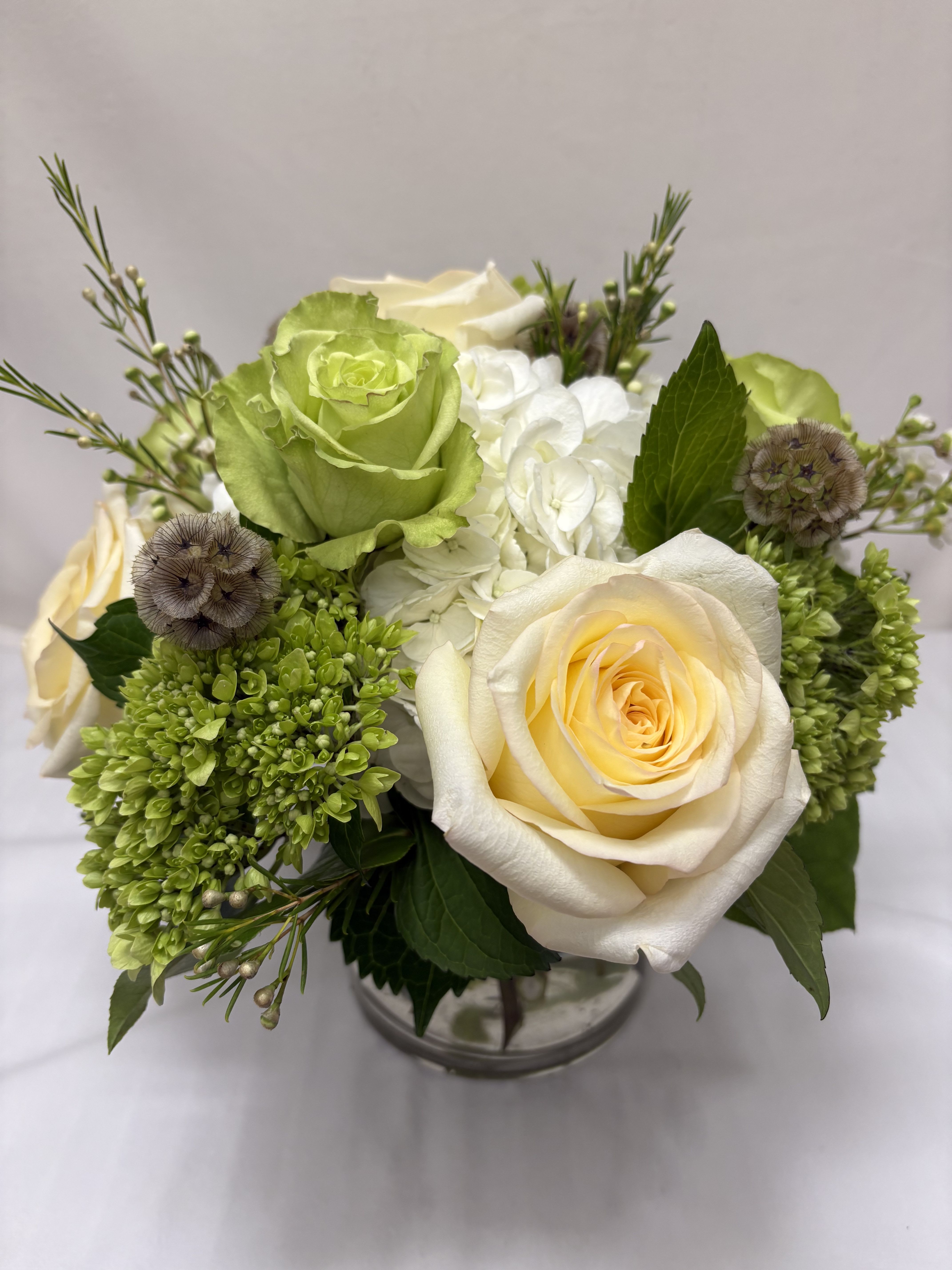 Compact arrangement of ivory and green roses with white hydrangea in a clear glass vase