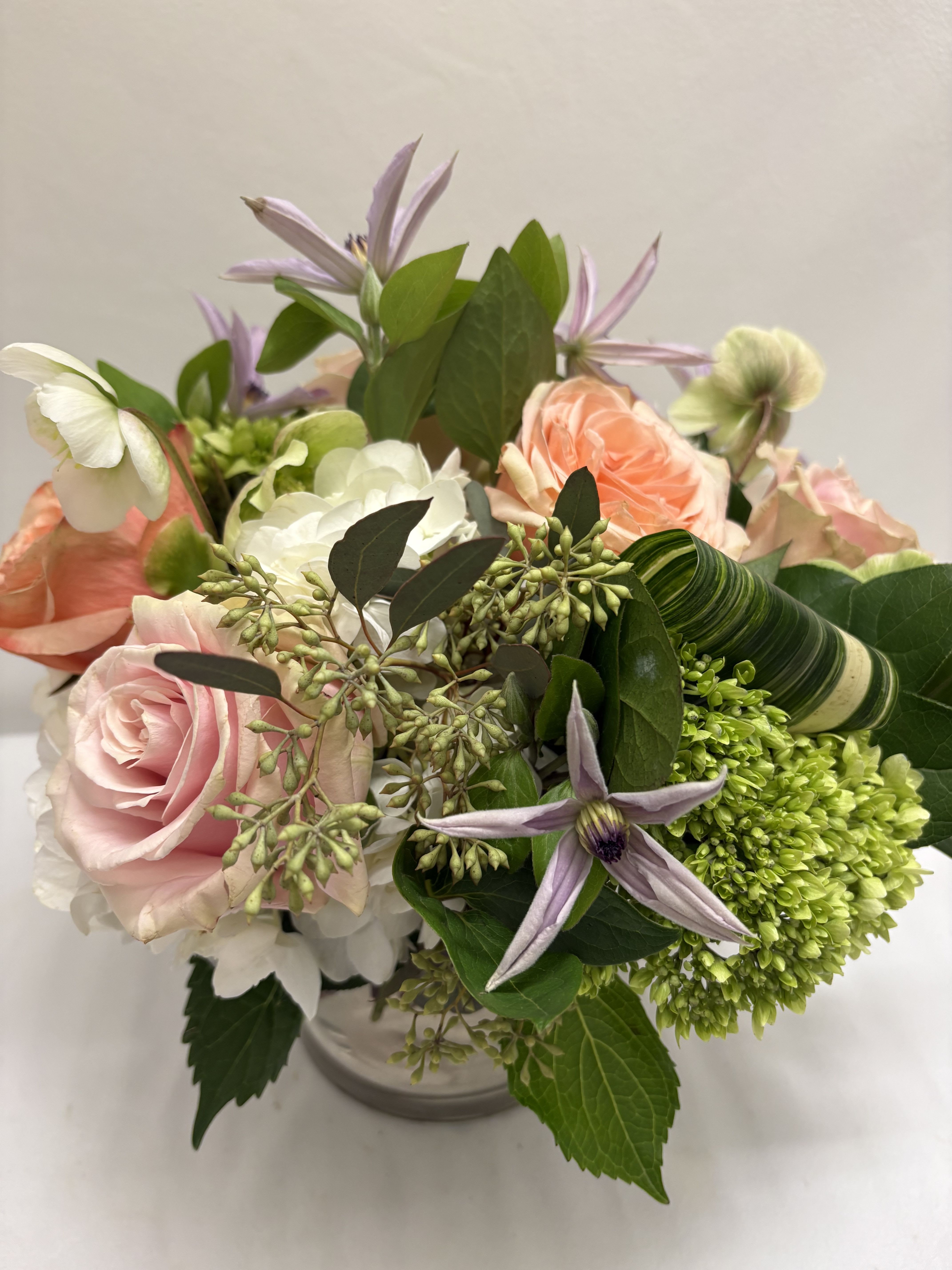 Short arrangement of peach and pink roses with white and green blooms in a clear glass vase