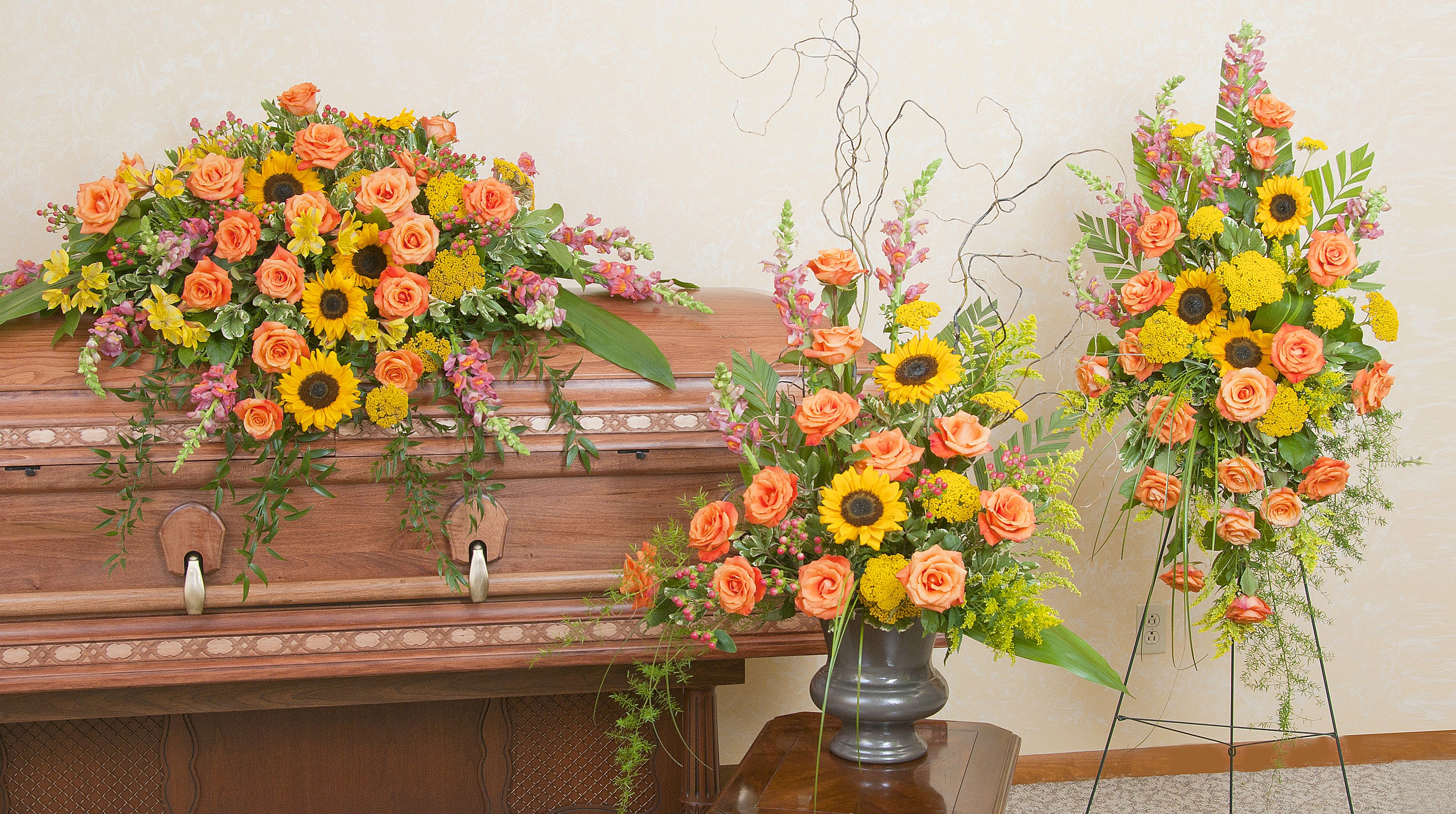 Three floral funeral arrangements with orange roses and sunflowers around a casket.