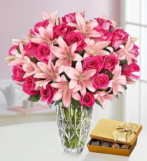 Pink roses and lilies in a glass vase beside a box of chocolates