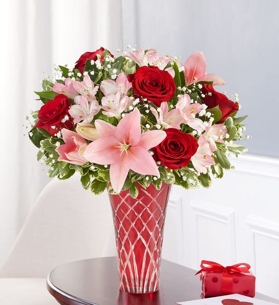 Red roses and pink lilies in a red glass vase with a gift box nearby