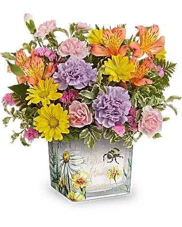  Floral Buzz Cube - Like a bee dancing through a hidden garden, this enchanting bouquet of orange, lavender, and yellow blooms in a vintage-inspired glass cube makes for a truly magical gift. This delightful arrangement features vibrant orange alstroemeria, lavender carnations, miniature pink carnations, yellow daisy spray chrysanthemums, and pink sinuata statice, all beautifully accented with leatherleaf fern and pitta negra. It is presented in Floral Buzz Cube, making it a charming addition to any tablescape.