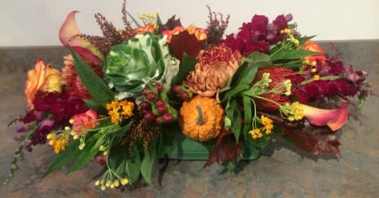 Low autumn centerpiece with orange calla lilies, burgundy flowers, kale and small gourds in a green container