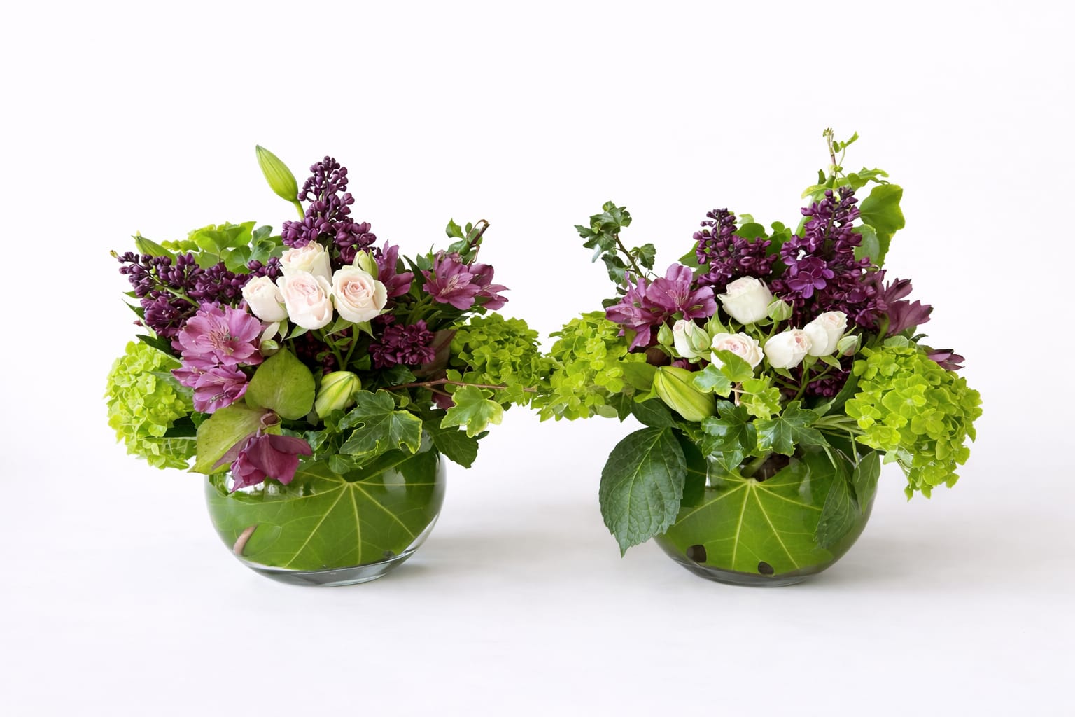 Twin Flames - A pair of mirrored, garden-style arrangements bursting with fresh spring color, each featuring vibrant green hydrangea, soft blush roses, and clusters of rich purple lilac. Accents of mauve alstroemeria and hellebore weave through glossy ivy and layered greenery, all artfully arranged in low glass bowls lined with bold green leaves, creating a lush, organic, and perfectly balanced duo.