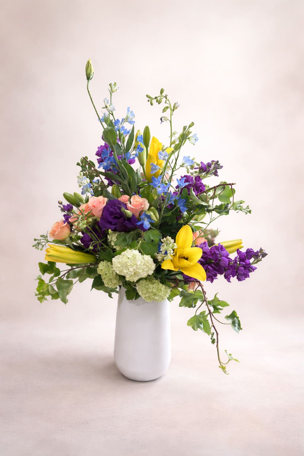 First Holy Communion - This arrangement has a light, garden-inspired feel, artfully composed in a smooth white ceramic vase that lets the colors take center stage. Bright yellow lilies open outward and upward, creating bold focal points among soft peach roses and rich purple blooms that add depth and contrast. Airy stems of delicate blue delphinium weave through the design, giving it movement and a slightly wild, just-gathered character. Rounded clusters of pale green hydrangea provide a soft base, while trailing ivy and varied greenery extend the silhouette, lending a natural, cascading elegance.