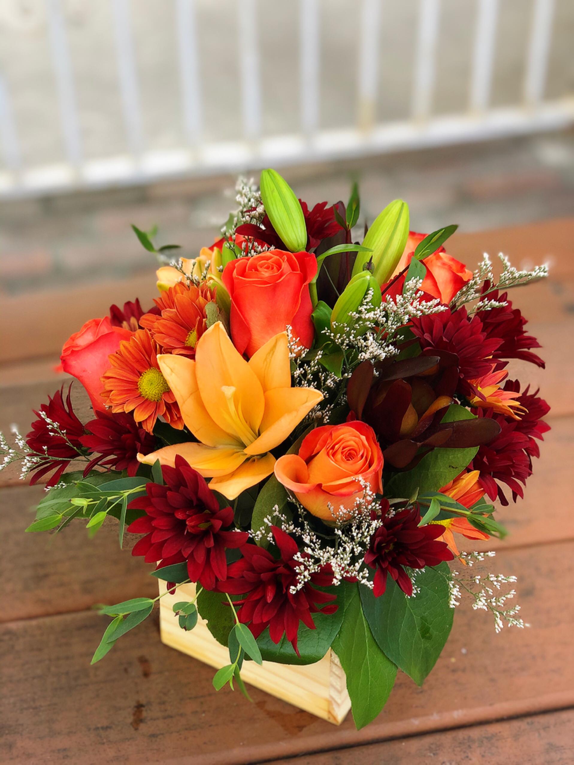 Fall in California  - -2 Stems Orange Lilies -6 Orange Roses -3 Red Daisies -2 Orange Daisies -White Filler -Greenery -4.75 Inches Wooden Square Vase