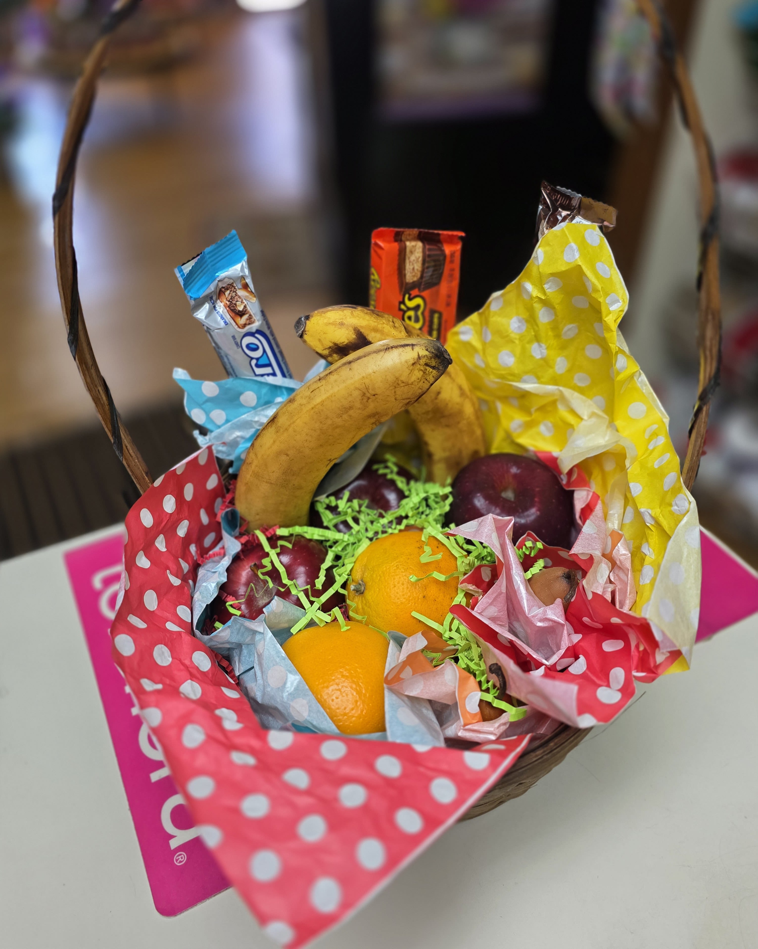 Gourmet Basket of Fruit &amp; Chocolate - Sunlit yellow bananas, glossy red apples, and juicy oranges spill from a woven basket alongside wrapped candy bars, making this fruit and chocolate gift basket a cheerful birthday basket or thank-you gift that feels bright and approachable. The playful polka dot tissue in red, yellow, blue, and pink gives the presentation a casual, celebratory energy-perfect for a coworker, friend, or someone you want to surprise with something comforting and fun.  Inside the basket you'll find a selection of seasonal fruit (bananas, oranges, apples) nestled on shredded paper filler with several wrapped candy bars for a sweet complement. Polka dot tissue paper frames the arrangement for a pop of color, and the sturdy woven basket makes a ready-to-gift presentation that's easy to carry and enjoy. Ideal for everyday gifting, birthdays, or a simple thank-you, this basket arrives assembled and ready to brighten someone's day.