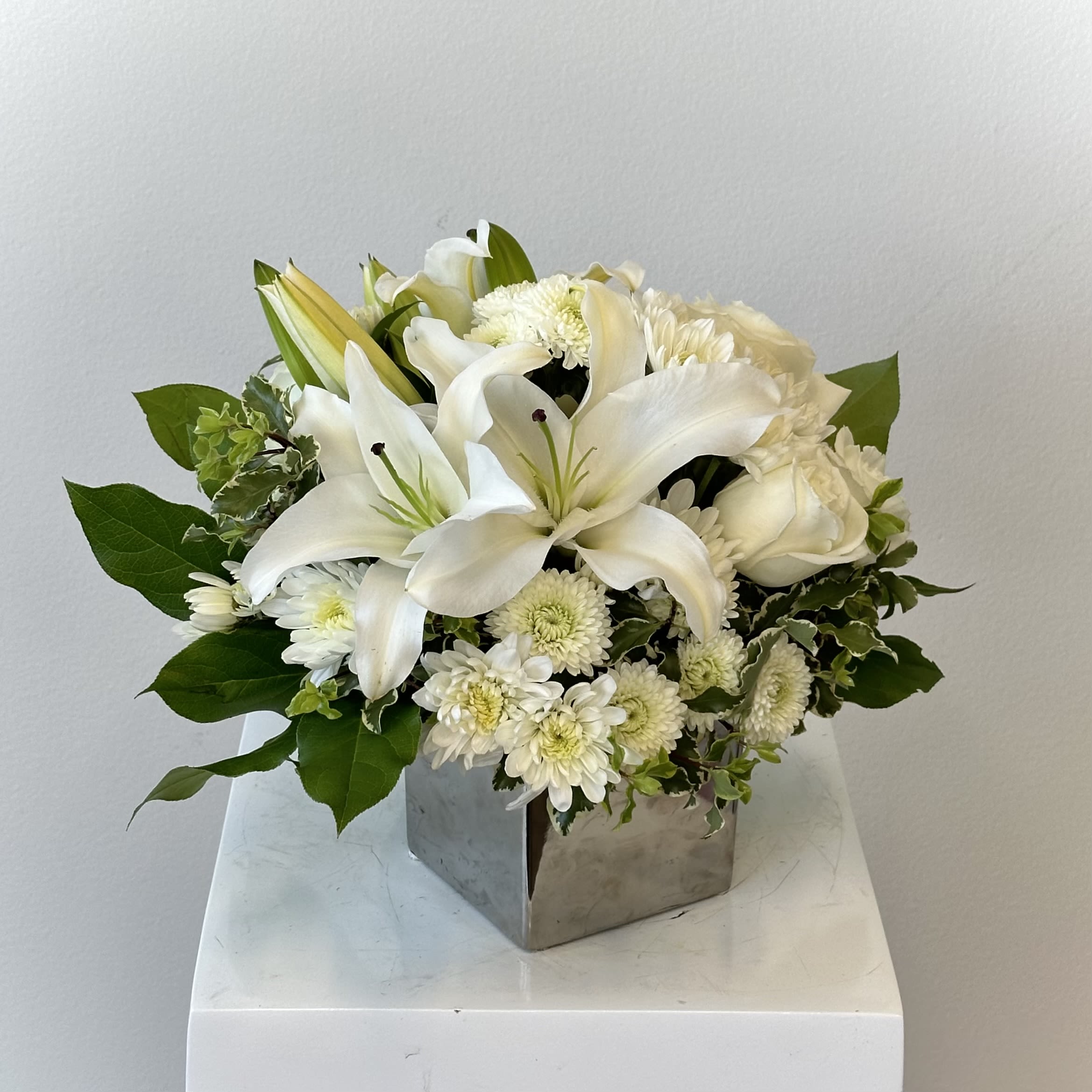 Peace and Joy Bouquet   - Celebrate serenity and heartfelt sentiment with the Peace and Joy Bouquet, a timeless all-white floral arrangement designed to bring comfort, elegance, and tranquility to any setting. Arranged in a modern cube vase, this beautiful all-around bouquet features a harmonious blend of fresh white lilies, white roses, white chrysanthemums, and delicate white carnations, accented with lush greenery for natural texture and balance. The classic white color palette symbolizes peace, remembrance, purity, and hope, making this arrangement a thoughtful choice for sympathy gifts, memorial services, celebrations of life, or simply to send a message of comfort and support.  Expertly designed to be viewed from every angle, the Peace and Joy Bouquet is a full, balanced arrangement that looks stunning on a dining table, memorial display, office desk, or entryway. Standing approximately 10 inches tall and 10 inches wide, it is the perfect size to make a graceful statement without overwhelming a space. The elegant cube vase adds a modern touch while highlighting the soft beauty of the blooms.  Whether you are honoring a loved one, offering condolences, or sharing a peaceful gesture of care, the Peace and Joy Bouquet delivers a meaningful expression of sympathy and grace. Fresh, classic, and thoughtfully arranged, it brings a calming presence wherever it is displayed.