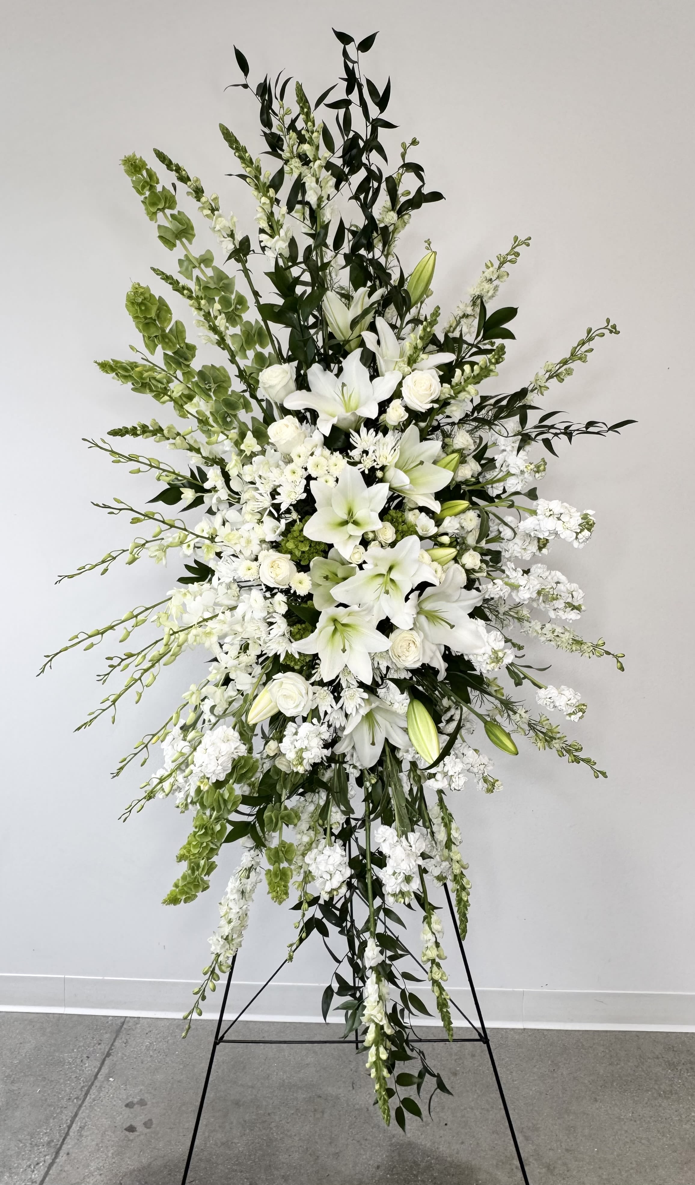 Grand Farewell - Grand Farewell is an extraordinary sympathy spray designed to create a breathtaking and unforgettable tribute in honor of a loved one. Standing approximately 7ft tall and 3.5ft wide, this grand floral display offers an elegant expression of peace, remembrance, and heartfelt sympathy for funerals, memorial services, celebrations of life, and graveside ceremonies. Featuring an all-white palette of premium fresh flowers, Grand Farewell combines timeless beauty with graceful sophistication to provide comfort and honor during life’s most difficult moments.  This luxurious arrangement includes fragrant white lilies, elegant white stock, towering white snapdragons, lush white roses, striking Bells of Ireland, and delicate dendrobium orchid buds. The crisp white blooms symbolize purity, peace, reverence, and eternal love, while the vibrant green Bells of Ireland add height, texture, and a symbol of hope and good fortune. Together, these flowers create a dramatic and serene presentation that beautifully reflects a life remembered with love and admiration.  Displayed on a standing easel, Grand Farewell makes a stunning focal piece suitable for funeral homes, churches, memorial chapels, and large services. Each arrangement is handcrafted by our florists using premium flowers to ensure every tribute is both visually impactful and deeply meaningful. Grand Farewell serves as a powerful expression of sympathy, respect, and everlasting remembrance for someone who will always be cherished.
