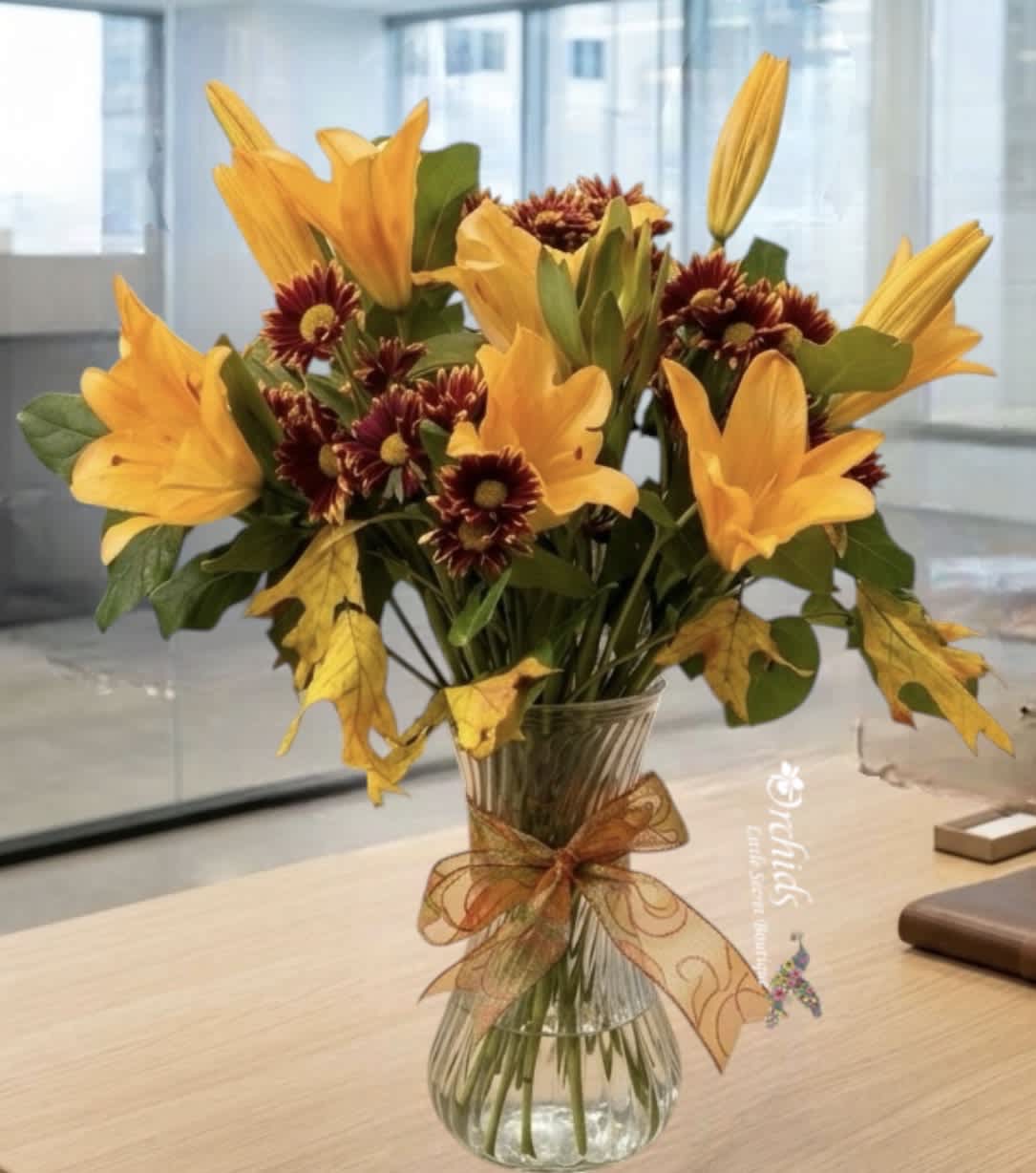 Orange lilies and burgundy daisies in a clear glass vase with a ribbon