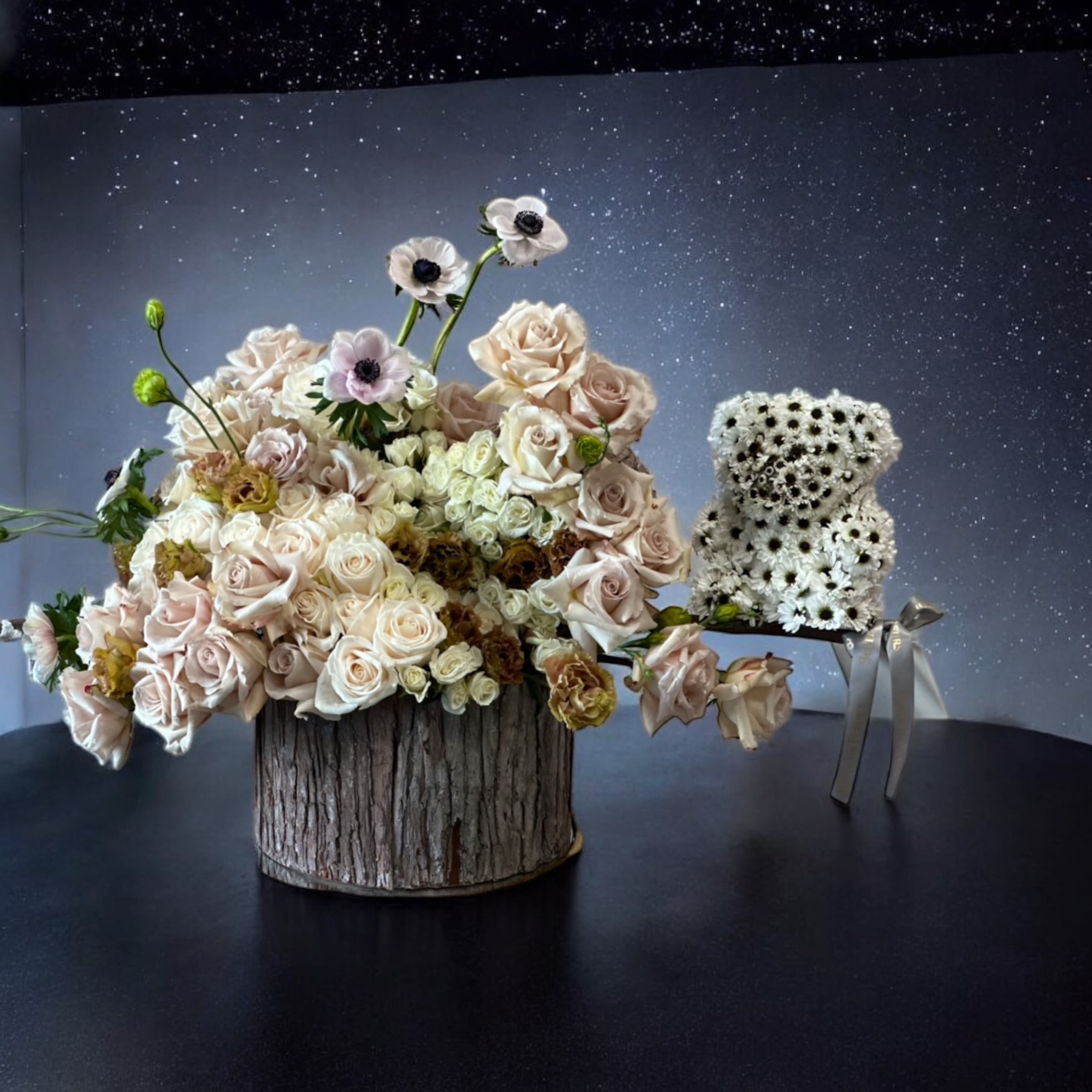 Blush roses and white anemones arranged in a rustic wood container beside a small white flower bear.