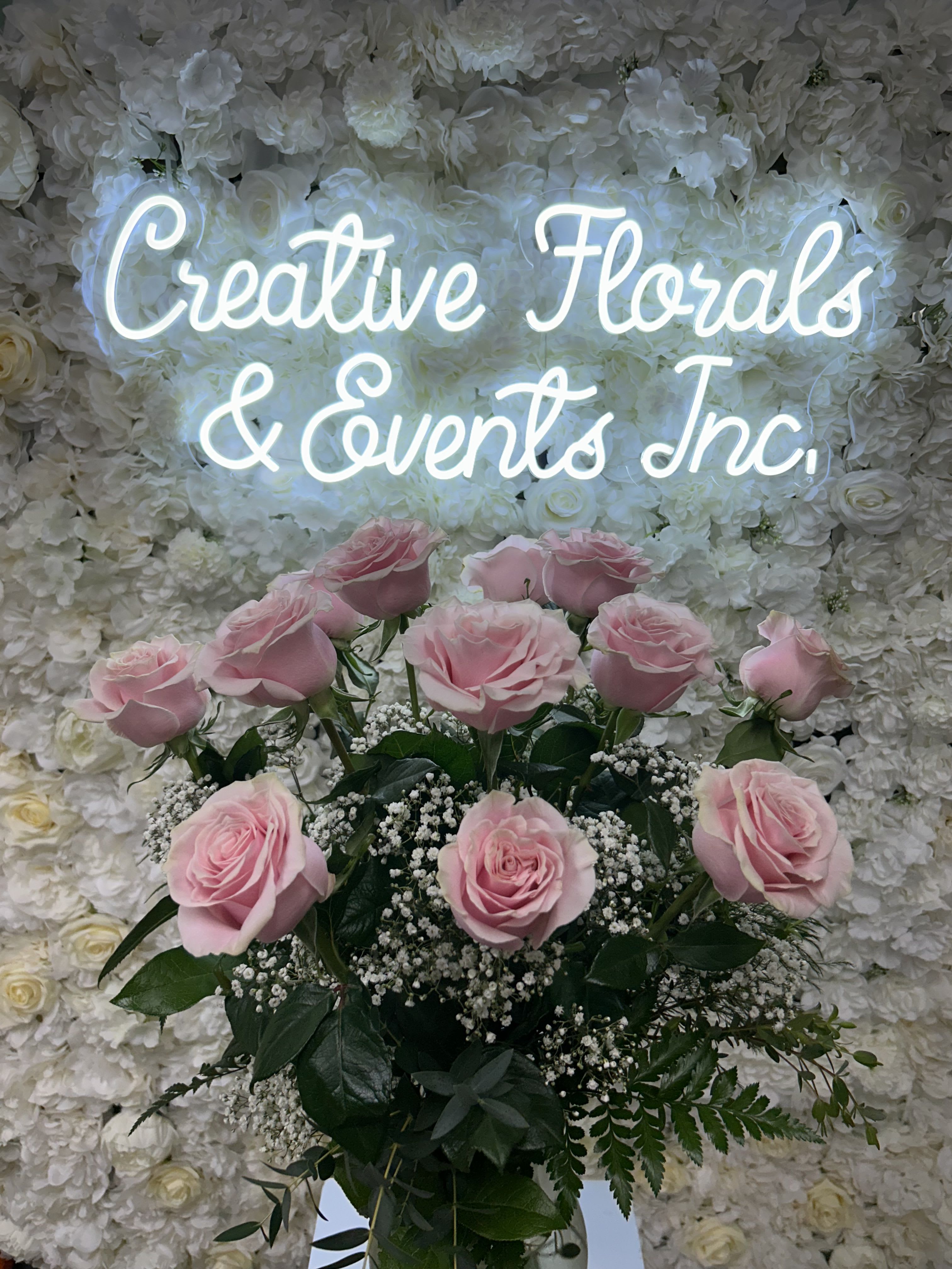 Pink roses arranged in front of a white floral wall with a glowing sign.