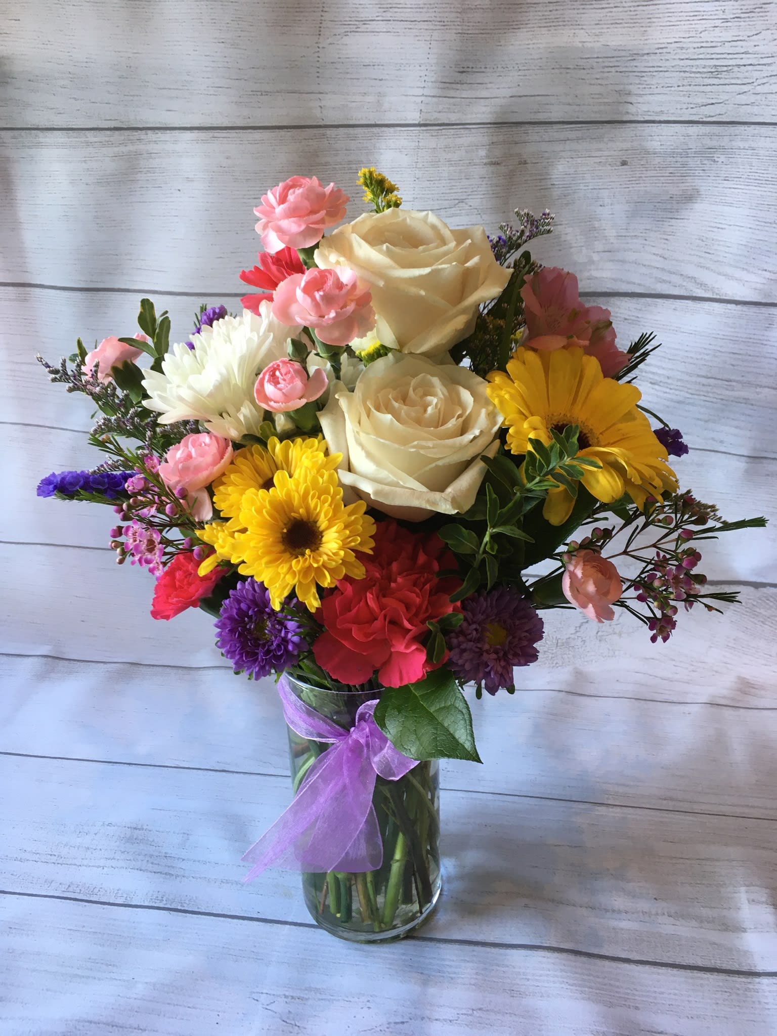 Mixed bouquet of roses, daisies, and carnations in a glass vase with a purple ribbon