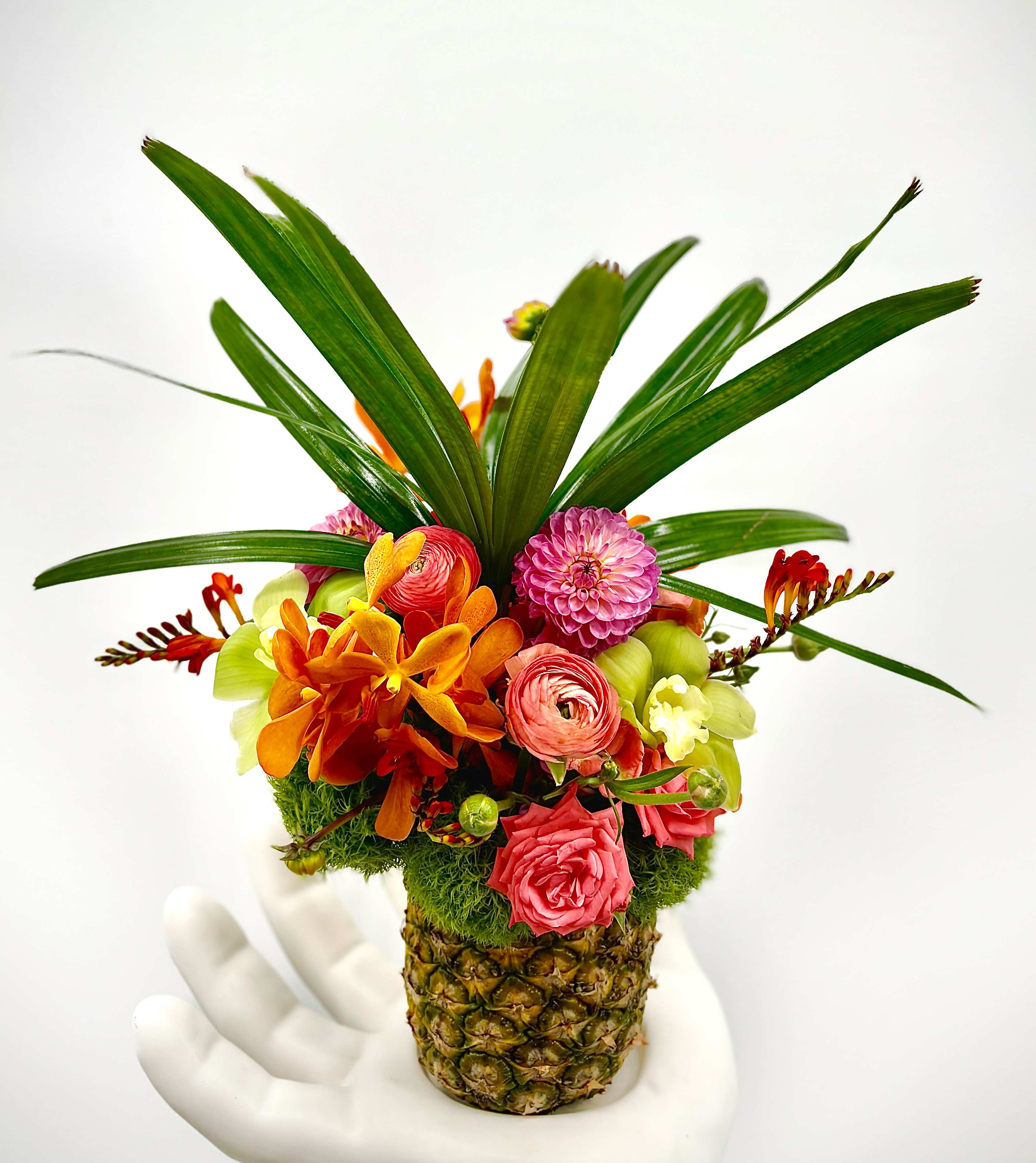 Lucas - This vibrant floral arrangement is set in a charming pineapple evoking tropical vibes. The bouquet features a lively mix of colorful flowers including orange orchids, pink ranunculus, bright dahlias, and lush green foliage. The greenery at the top mimics the leaves of a pineapple, adding to the tropical aesthetic. Perfect for parties and is the symbol of hospitality all over the South.