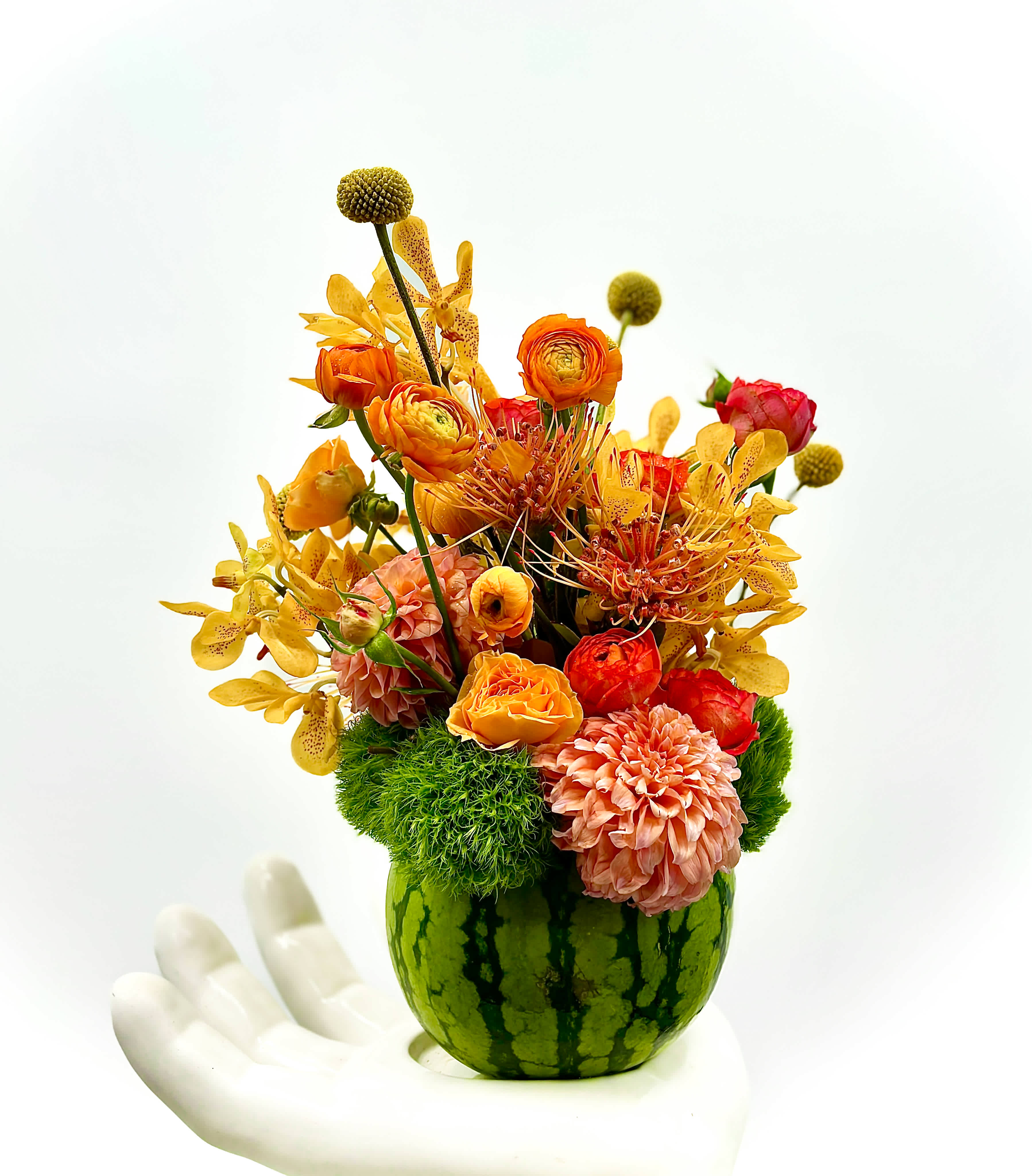 Ethan - This floral arrangement features a delightful mix of vibrant flowers in warm tones of orange, yellow, and pink, artfully displayed in a small watermelon  The composition includes cheerful marigolds, elegant orchids, and lush dahlias, complemented by pops of greenery.  This eye-catching display is perfect for summer gatherings, adding a refreshing and lively touch to any setting.