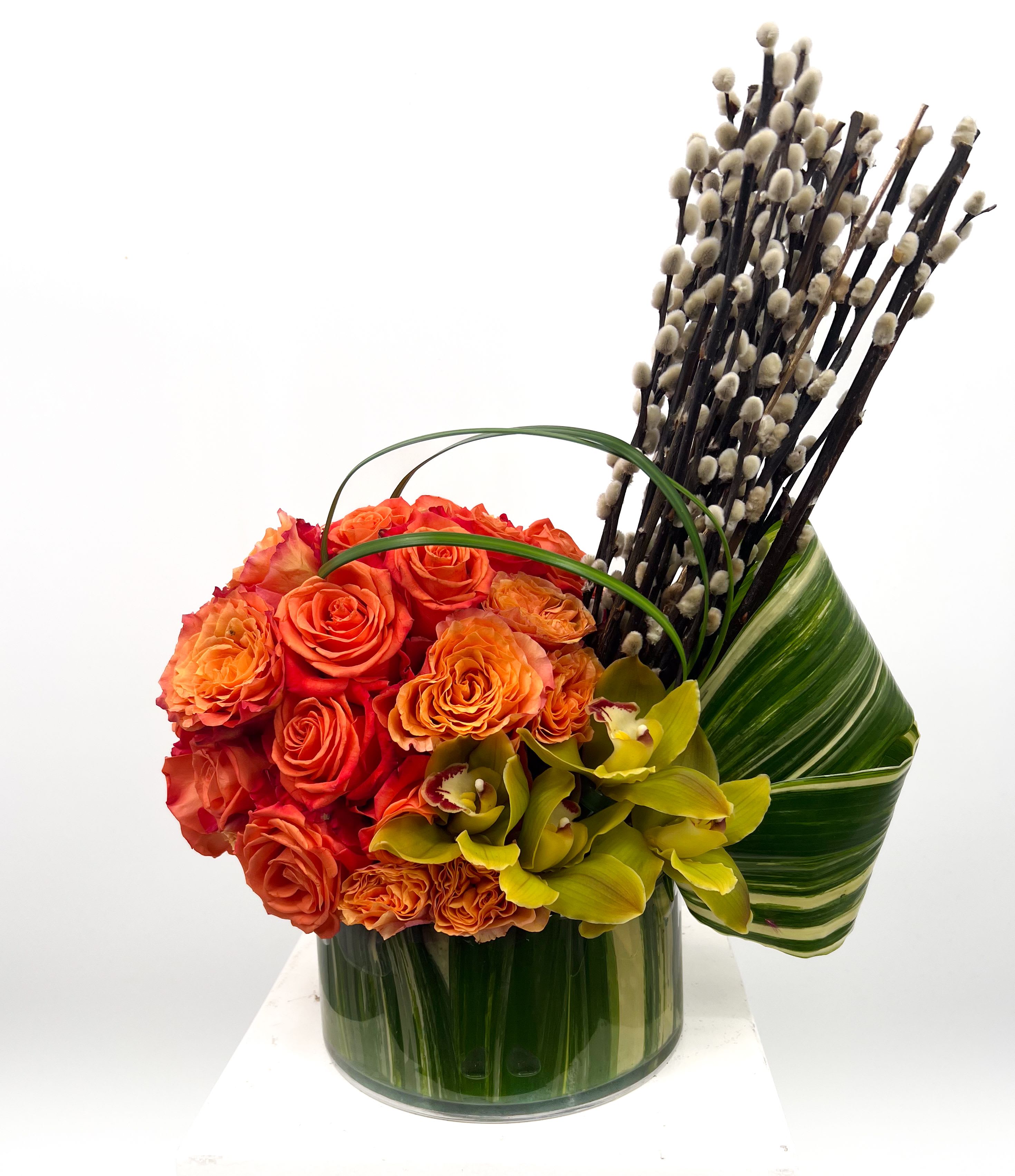 Harris - This vibrant floral arrangement features a stunning bouquet of fiery orange roses complemented by lush green leaves and delicate yellow-green orchids. The arrangement is further accentuated by an artistic addition of tall, slender pussy willow branches, creating a dynamic and elegant display. The flowers are neatly arranged in a clear cylindrical vase, making it a perfect centerpiece for any occasion.