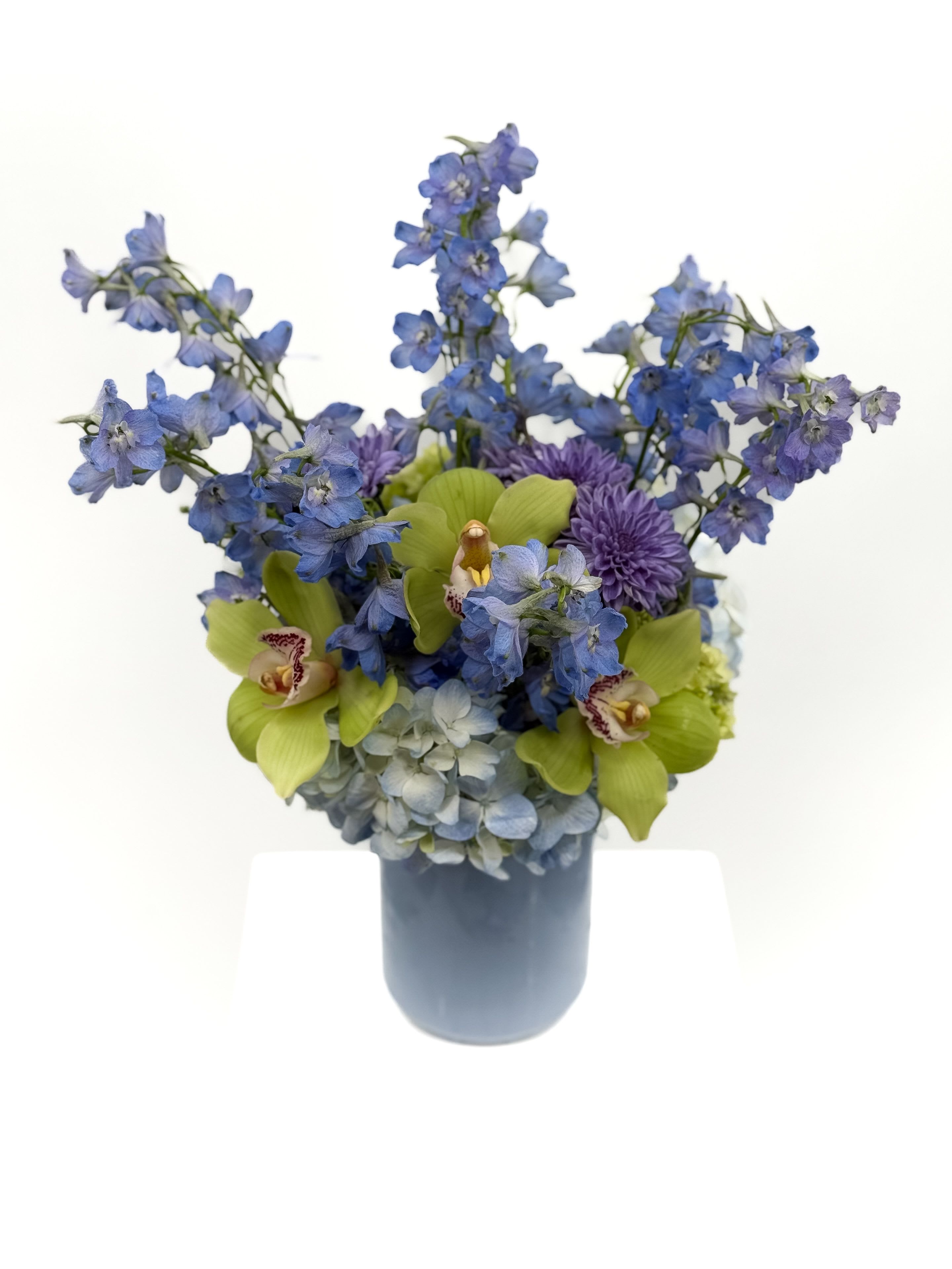 Peri - This artful arrangement showcases soothing periwinkle delphinium paired with lush hydrangeas, vibrant green cymbidium orchids, and soft lavender mums. Set in a sleek blue vase, it’s a dreamy blend of color and texture—perfect for thoughtful gifting, elegant events, or simply bringing calm luxury into any space.