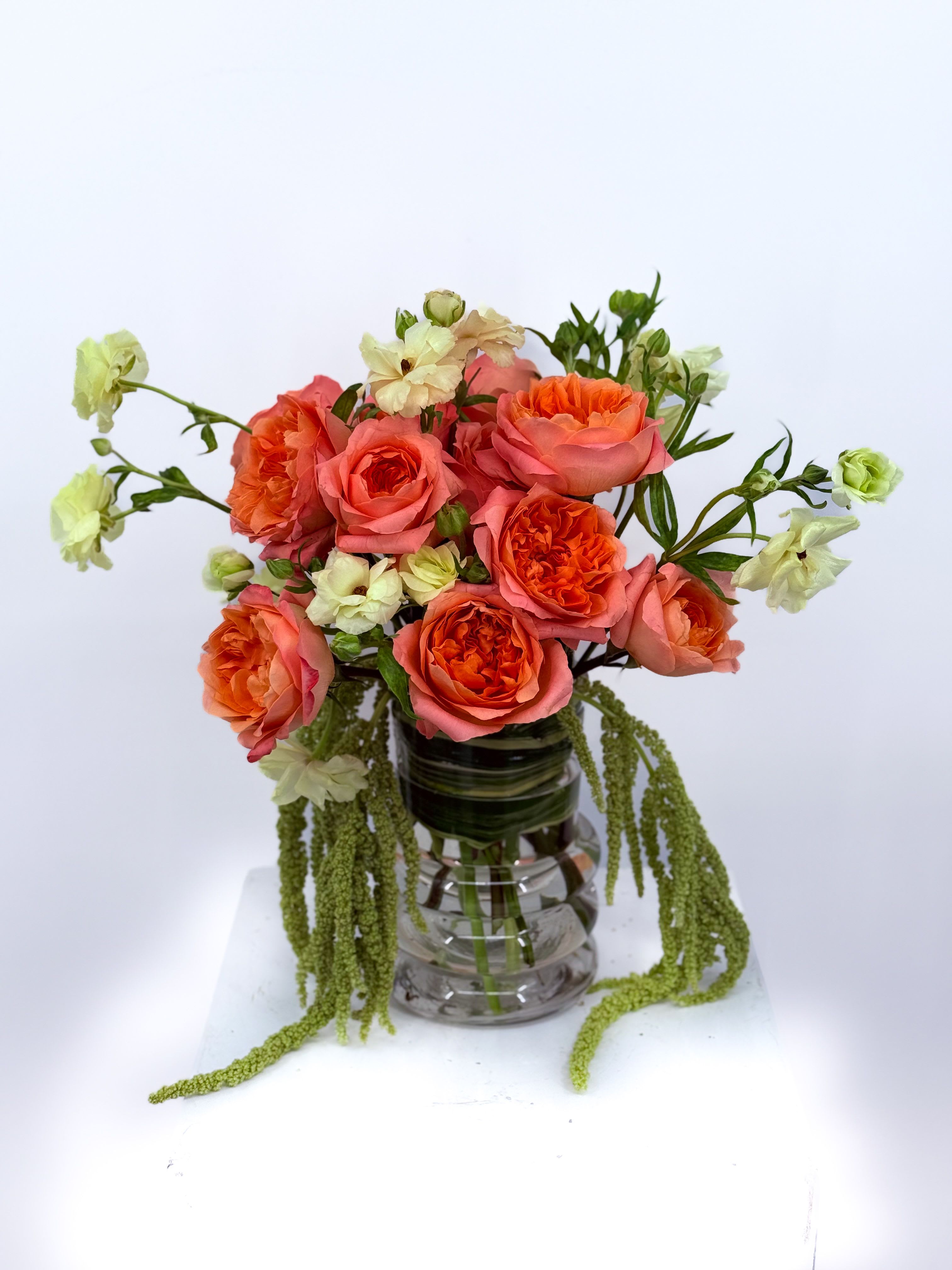 Jenny - Bursting with vibrant charm, this bouquet of coral garden roses and soft cream blooms brings pure joy to any space. The lush roses steal the spotlight with their ruffled petals, while cascading greenery adds an elegant, flowing touch. Perfect for brightening someone’s day, celebrating a special moment, or simply treating yourself to a little luxury.