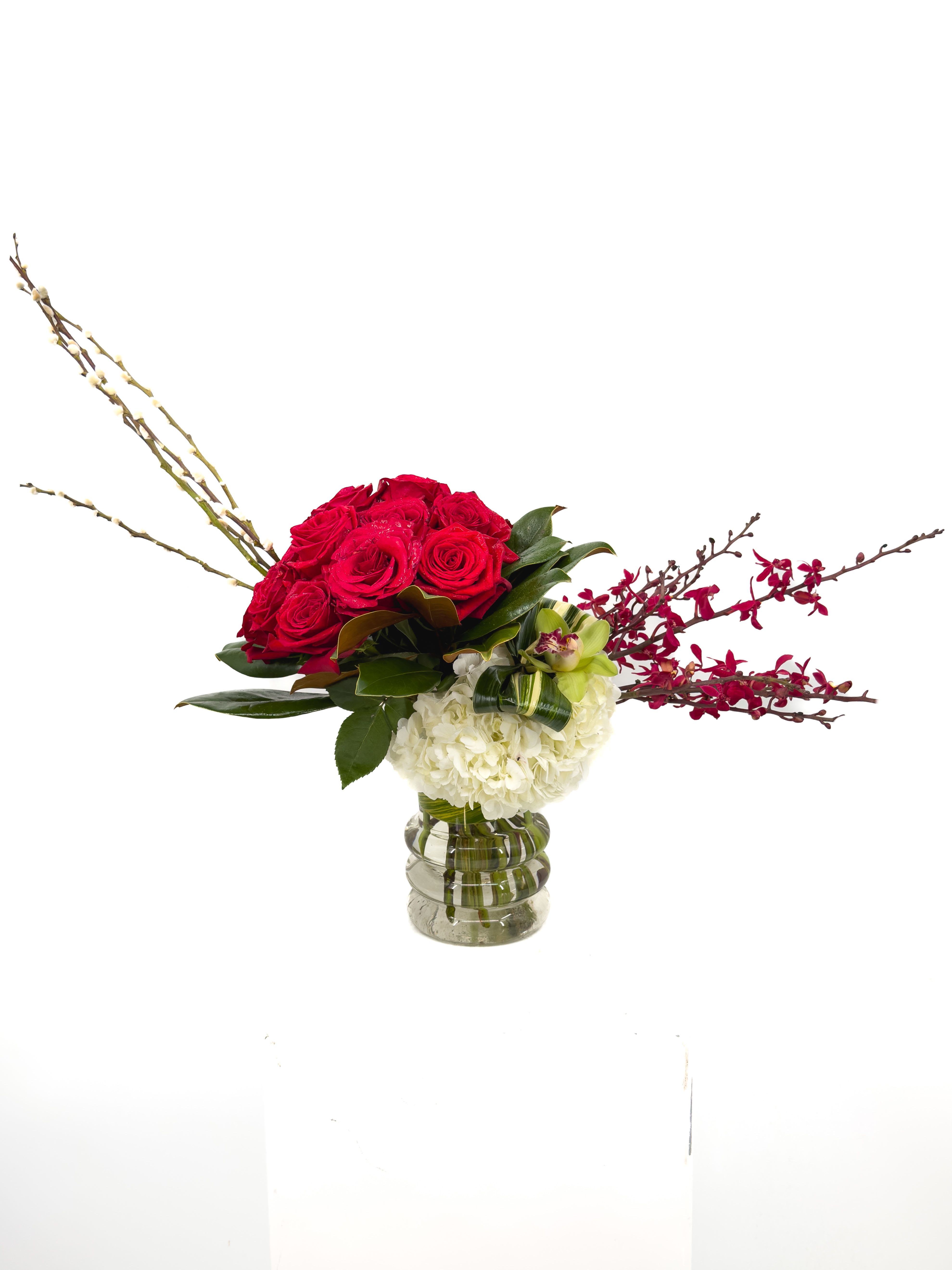Tara - Striking yet effortlessly elegant, this floral arrangement is a sophisticated expression of love and style. A lush Texas dozen* of velvety red roses takes center stage, symbolizing passion and devotion, beautifully balanced by soft white hydrangea that adds volume and grace. Delicate orchid accents and sculptural branches extend outward, creating a dramatic, contemporary silhouette with graceful movement. Designed in a clear glass vase, the arrangement feels light, architectural, and refined—perfect for modern interiors and elevated occasions. The contrast of bold reds, crisp whites, and fresh greenery creates a timeless palette that feels both powerful and romantic. Ideal for anniversaries, celebrations, or meaningful gestures, this arrangement makes a statement without saying a word.  Elegant. Confident. Unforgettable.   *Texas Dozen = 15