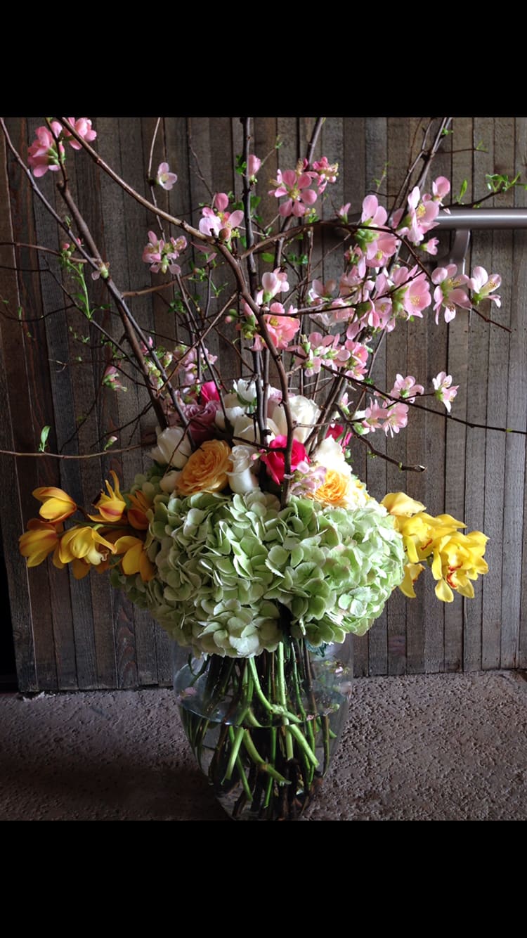 Tall floral arrangement with pink blossoms, hydrangeas, and yellow flowers in a glass vase