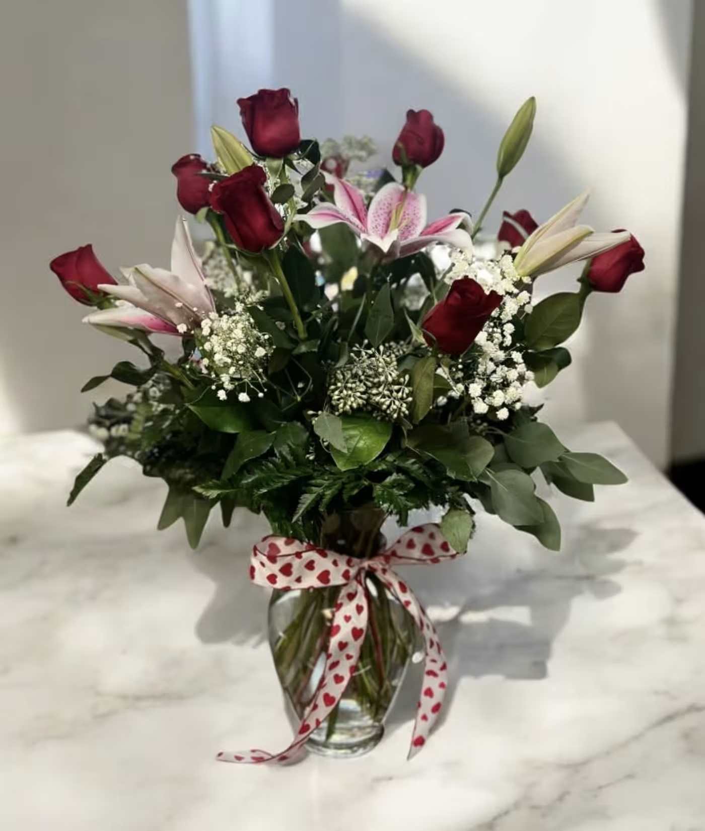 Bouquet of red roses and pink lilies in a glass vase with a heart ribbon