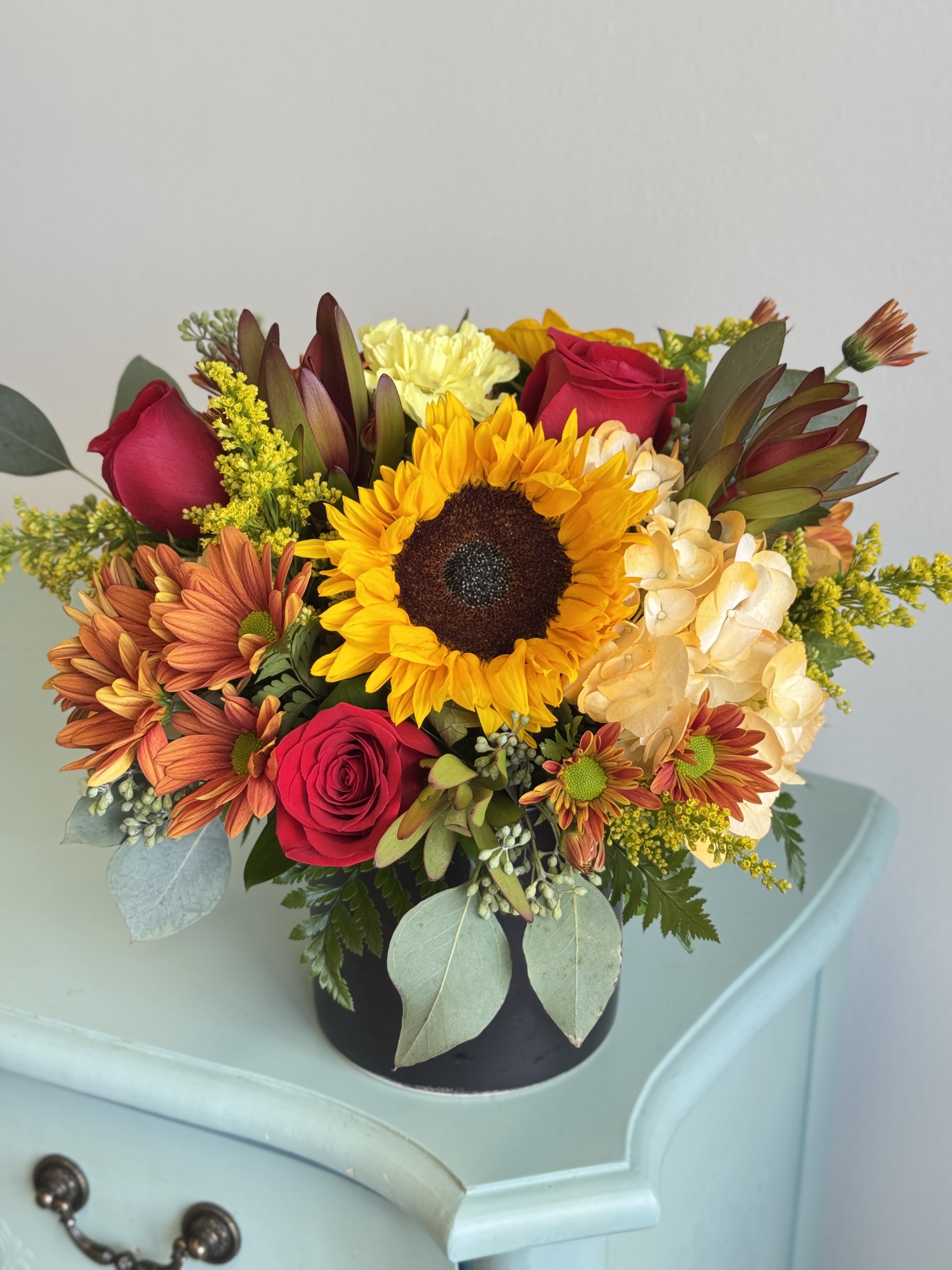 Mixed bouquet with a sunflower, red roses, and orange mums in a black vase