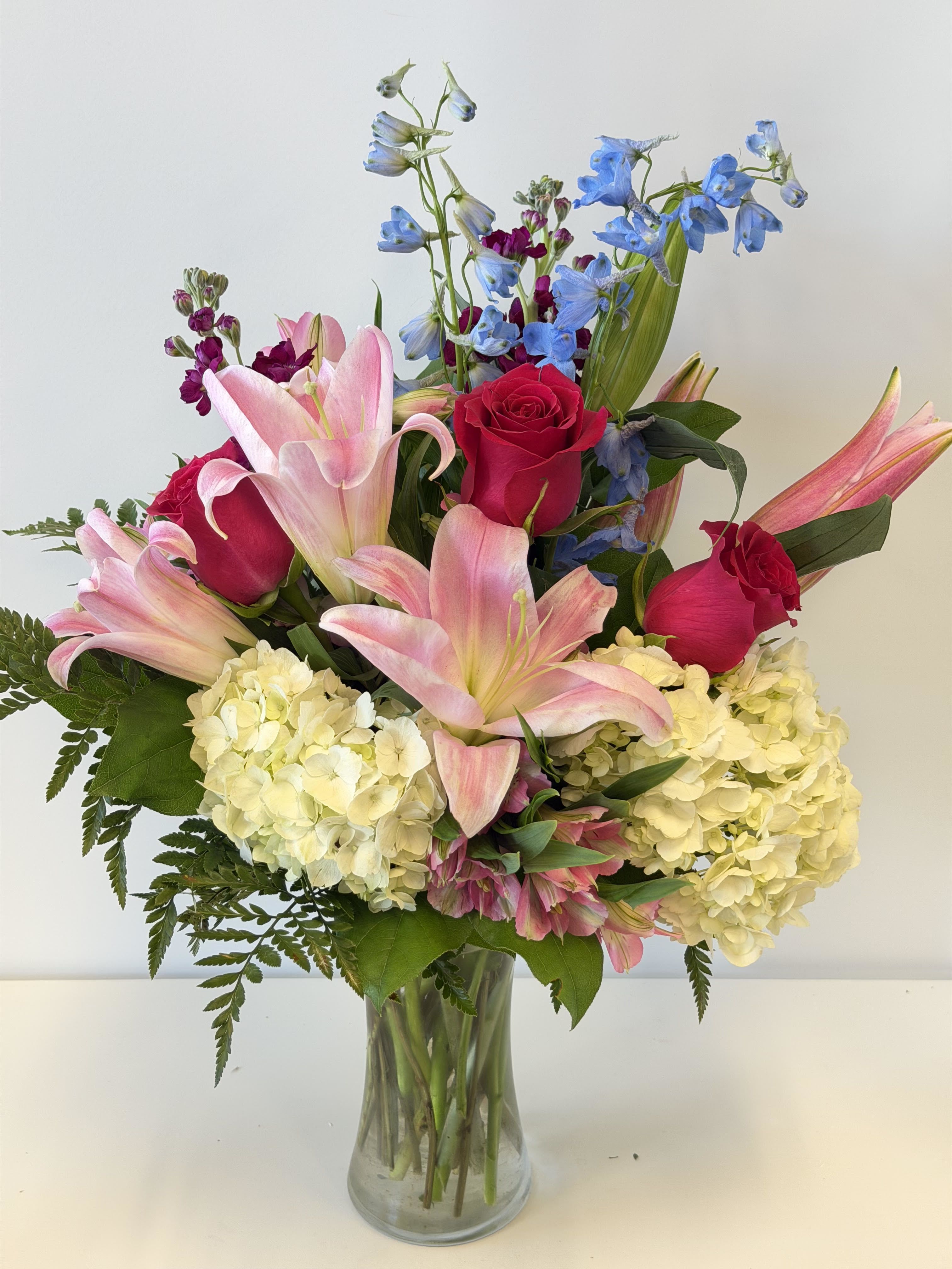 Bouquet of pink lilies, red roses, blue delphinium, and pale hydrangeas in a glass vase