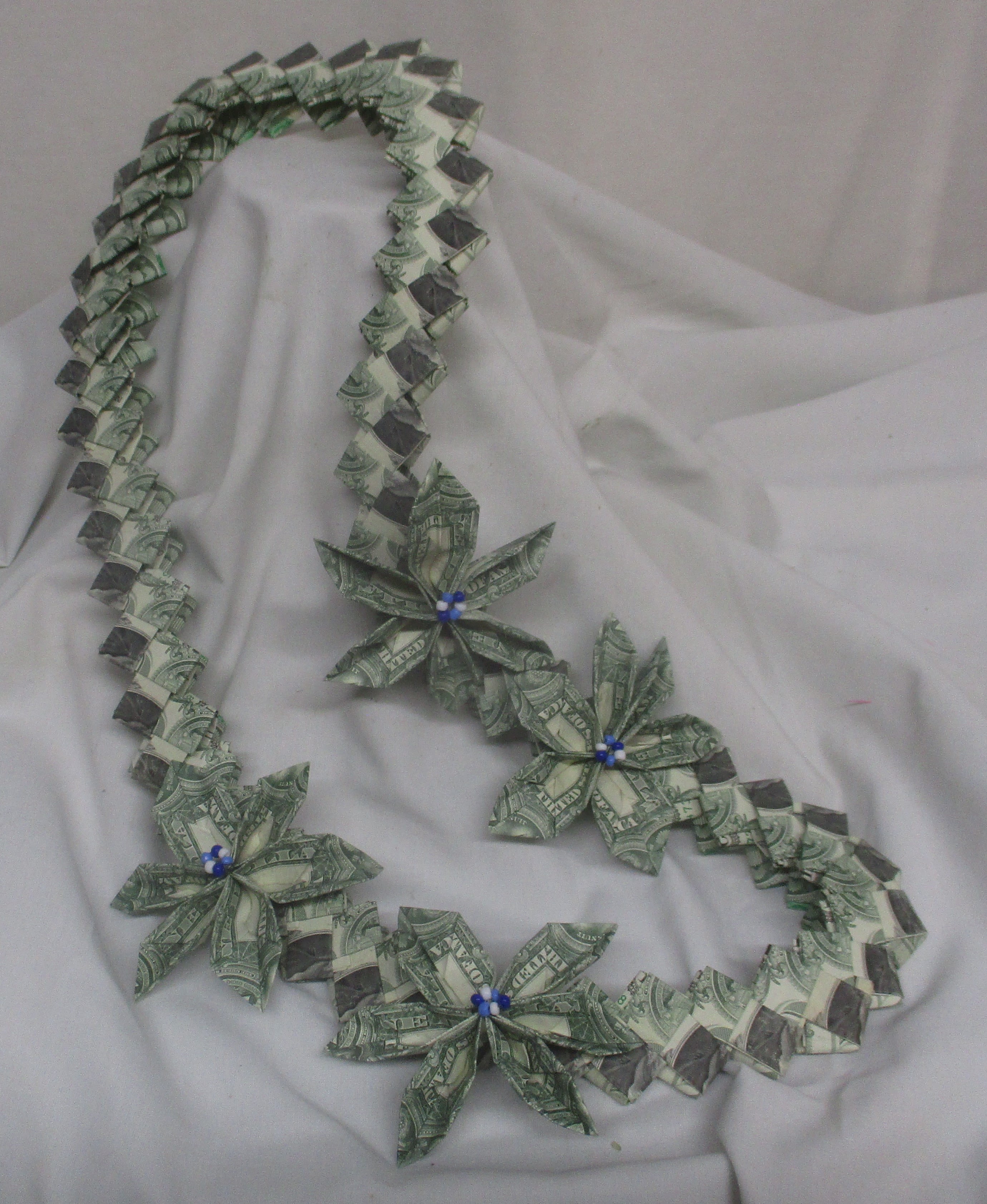 Lei - Lotus Money Lei - A bold, celebratory twist on a traditional gift-this green-and-white money lei turns graduation regalia into a keepsake the recipient will treasure. The folded dollar bills form lotus-style blooms along a loop, punctuated by small bead accents that lend a festive, ceremonial look perfect for honoring a new graduate or marking a major milestone.  Handcrafted with one-dollar bills arranged into a paper lei, the piece has a playful, handmade texture that reads as both thoughtful and fun. Lightweight and easy to wear over a gown, it arrives in a gift box, ready to drape for the ceremony. Ideal for graduations and congratulations, it's the kind of gift that makes the graduate feel celebrated and seen-order early while quantities last.