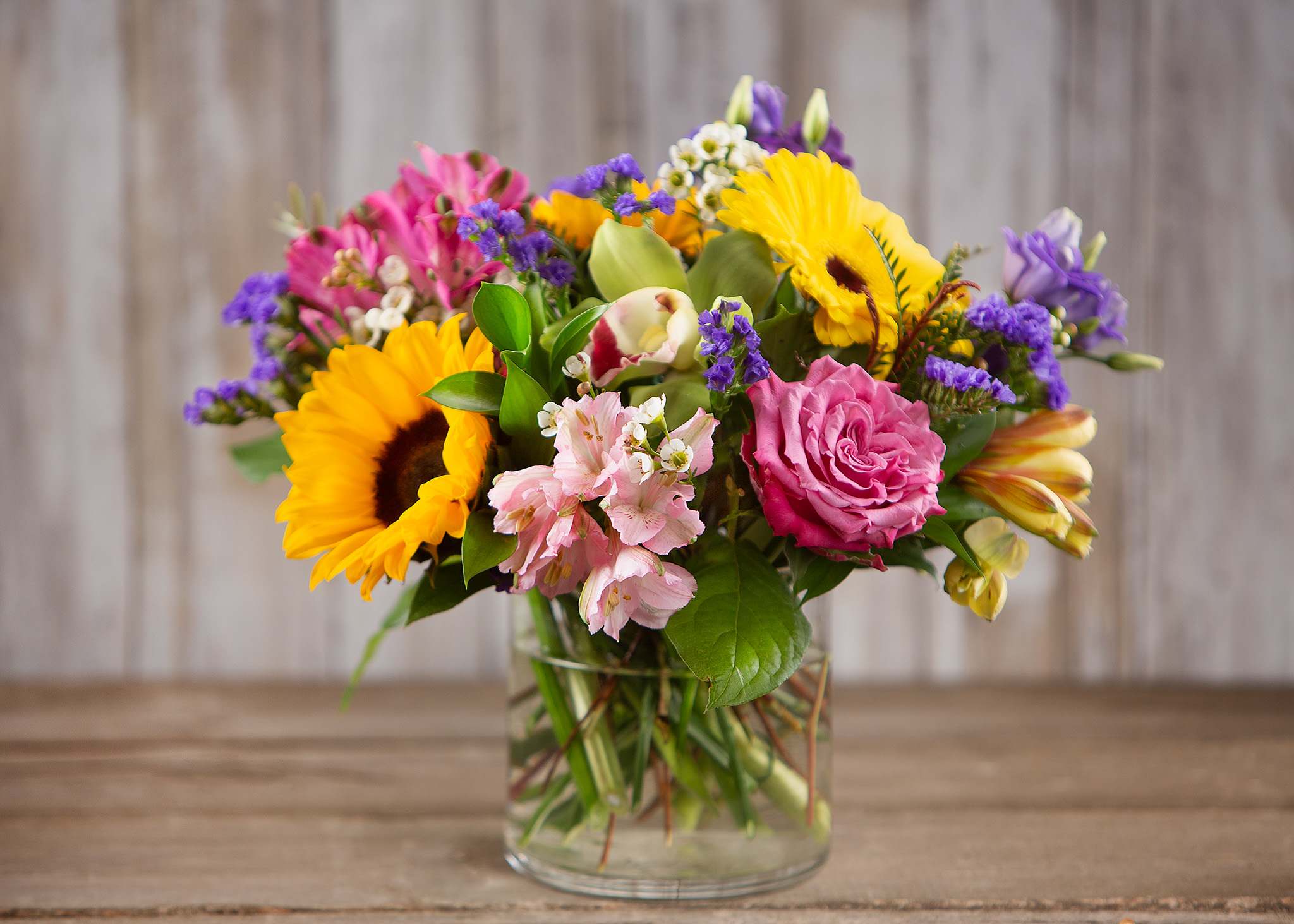 Mixed bouquet with sunflowers, pink roses, and purple blooms in a glass vase